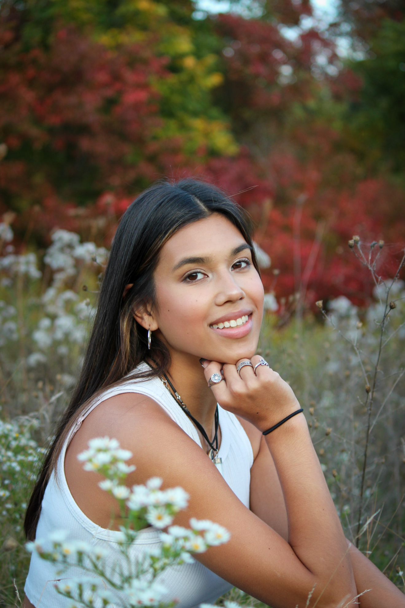 A young woman with long dark hair and light skin smiling outdoors among spring or summer flowers, with red and green autumn-colored trees in the background.