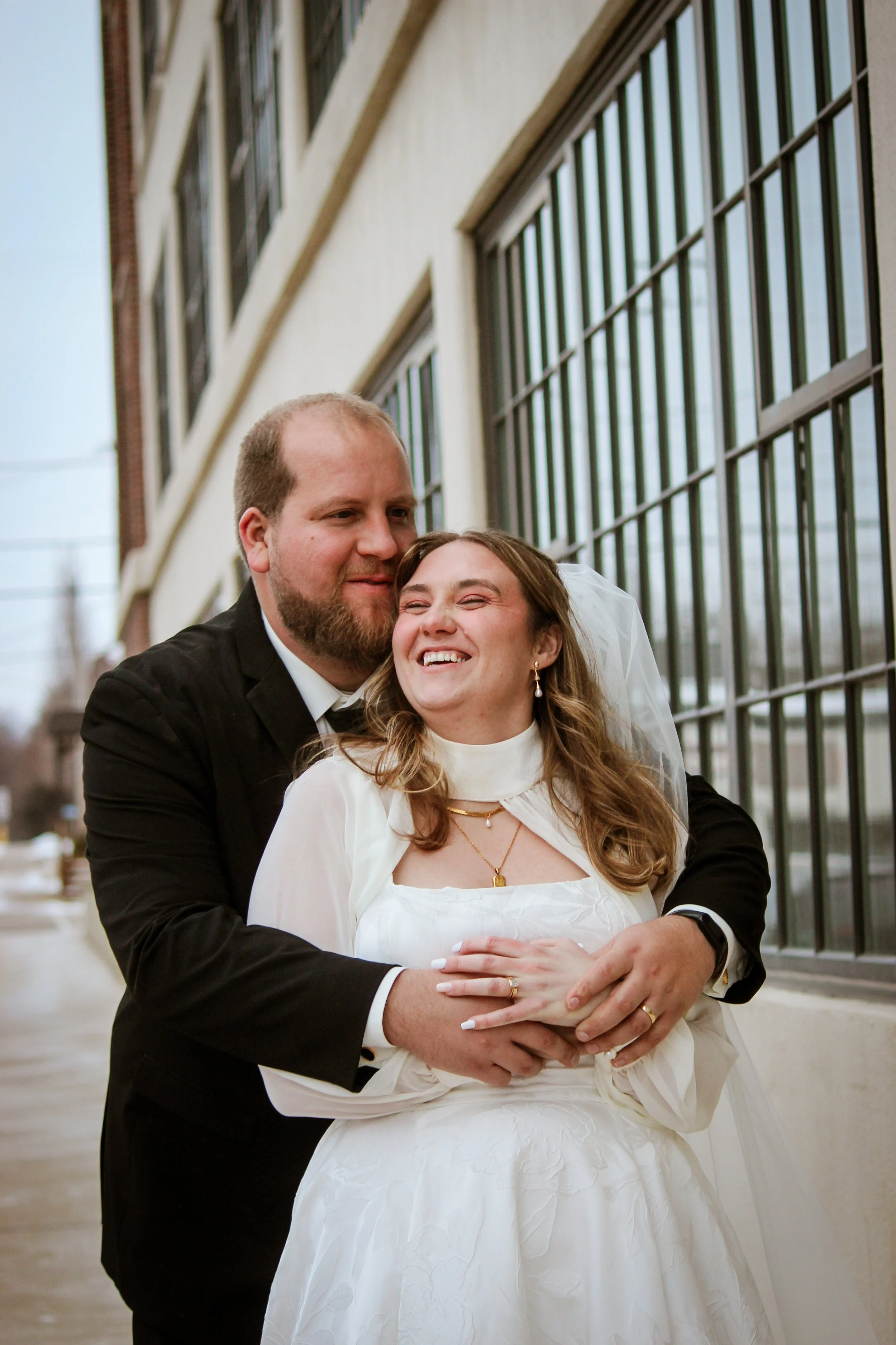A happy couple in wedding attire hugging outdoors near a building with large windows, celebrating their wedding day.