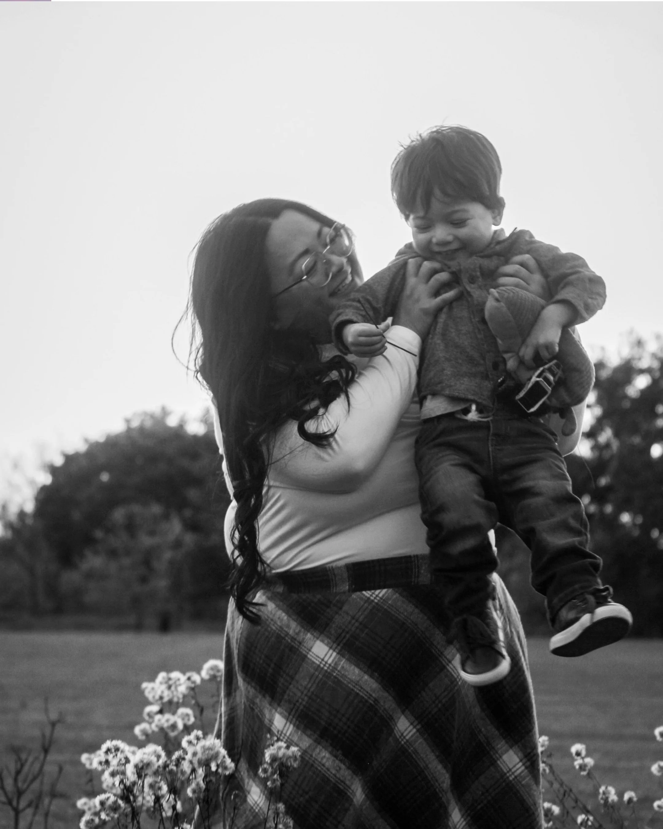 A woman with long, curly hair and glasses is lifting a young boy in the air outdoors, both smiling. The background features trees and flowers, suggesting a park or garden setting.