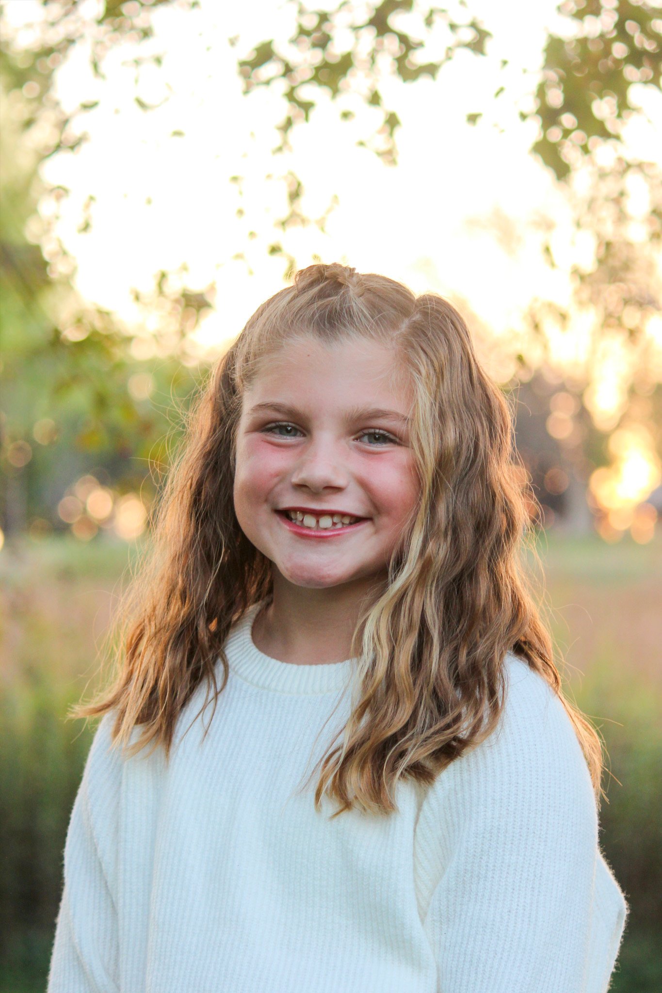 A young girl with long wavy blonde hair and a missing front tooth smiling outdoors at sunset, wearing a white sweater.