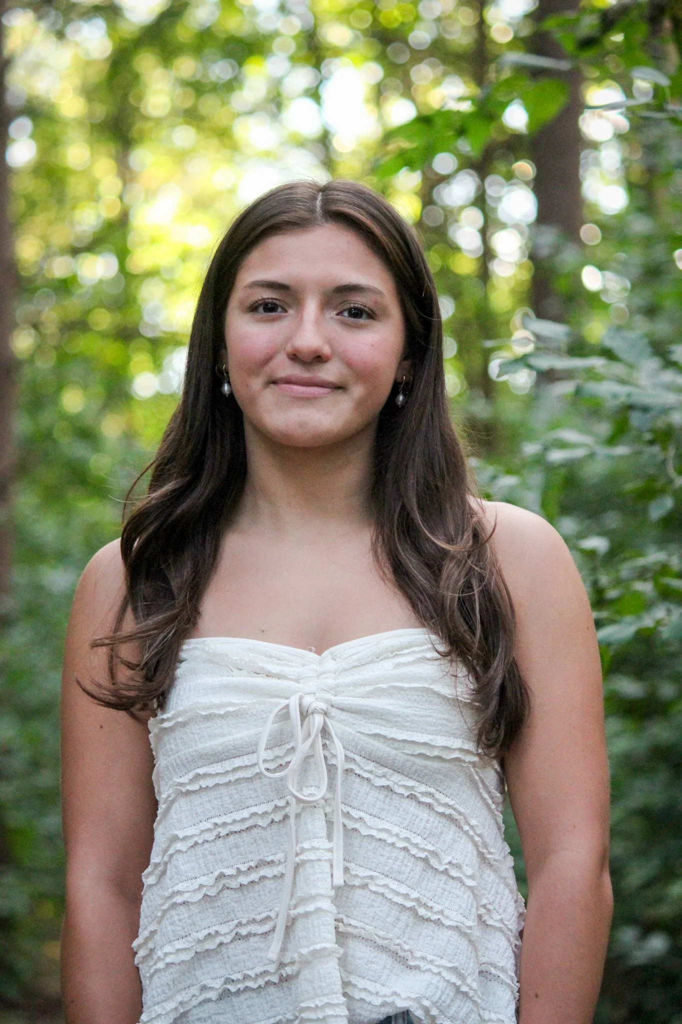 A young woman with long brown hair, wearing a white strapless top with a bow detail, standing in a lush green forest with sunlight filtering through the trees.