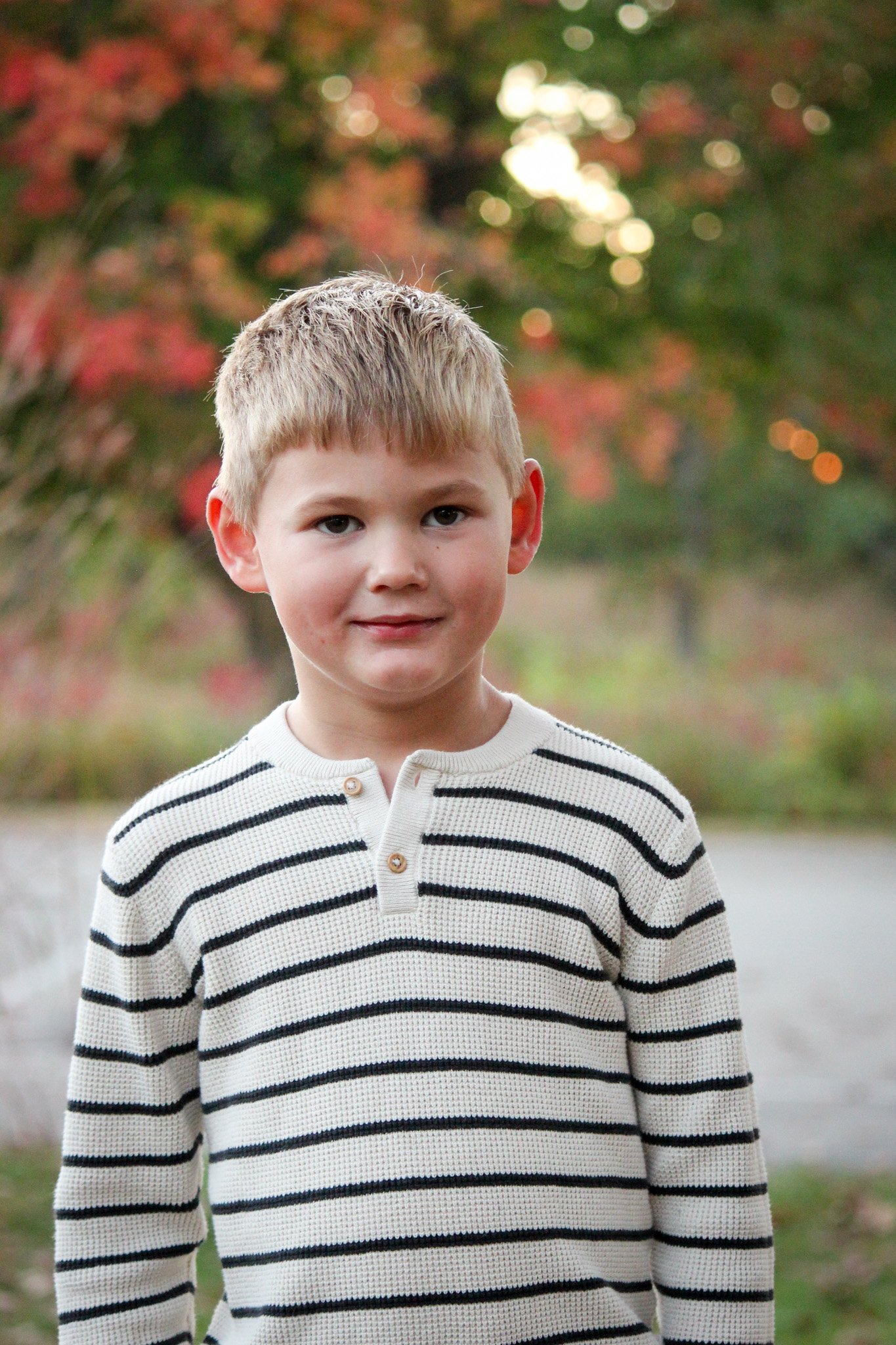 A young boy with blonde hair wearing a white and black striped sweater, standing outdoors with autumn trees and blurred foliage in the background.