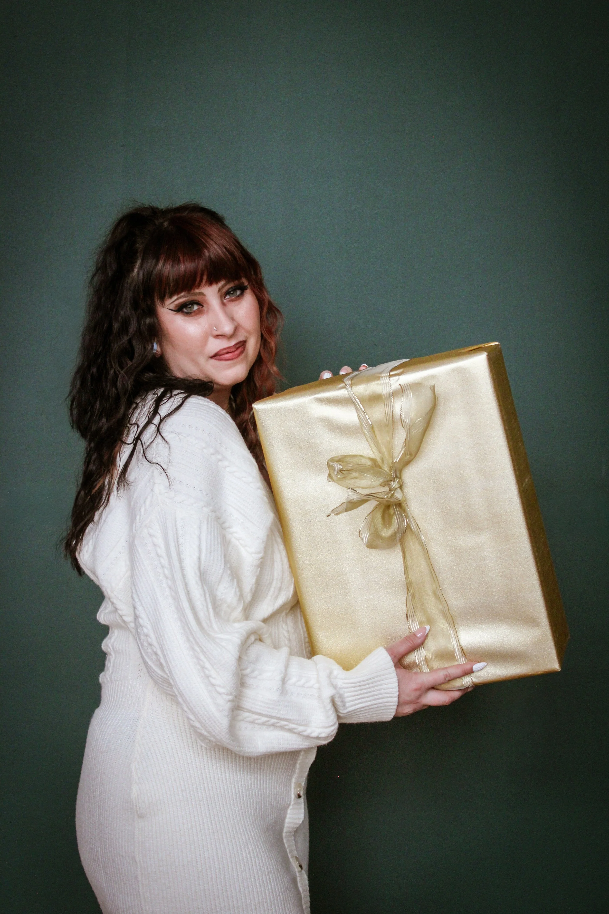A woman with wavy brown hair and a nose piercing holding a large, gold wrapped gift with a bow.