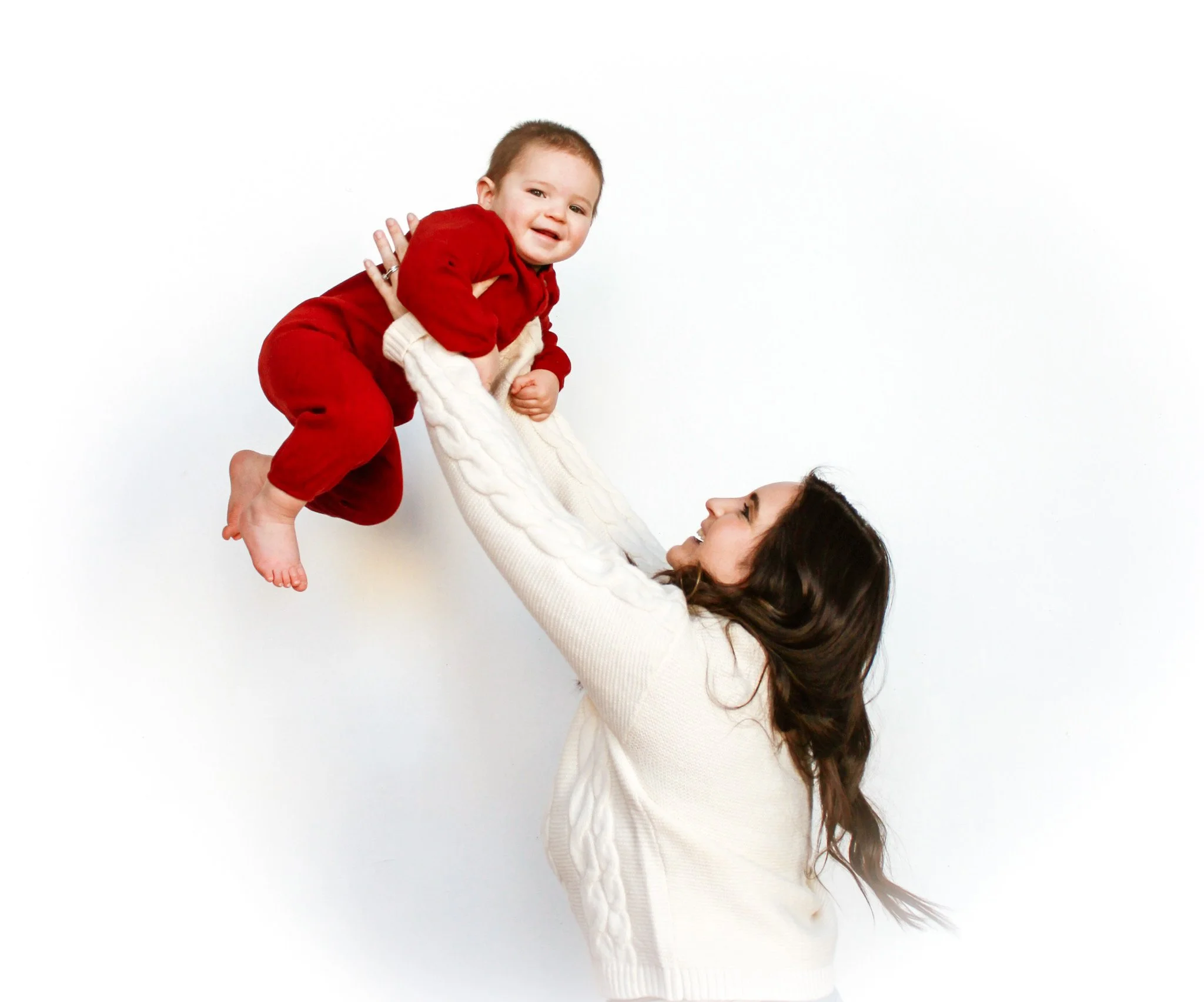 A joyful woman lifting a smiling young child in red clothes against a plain white background.