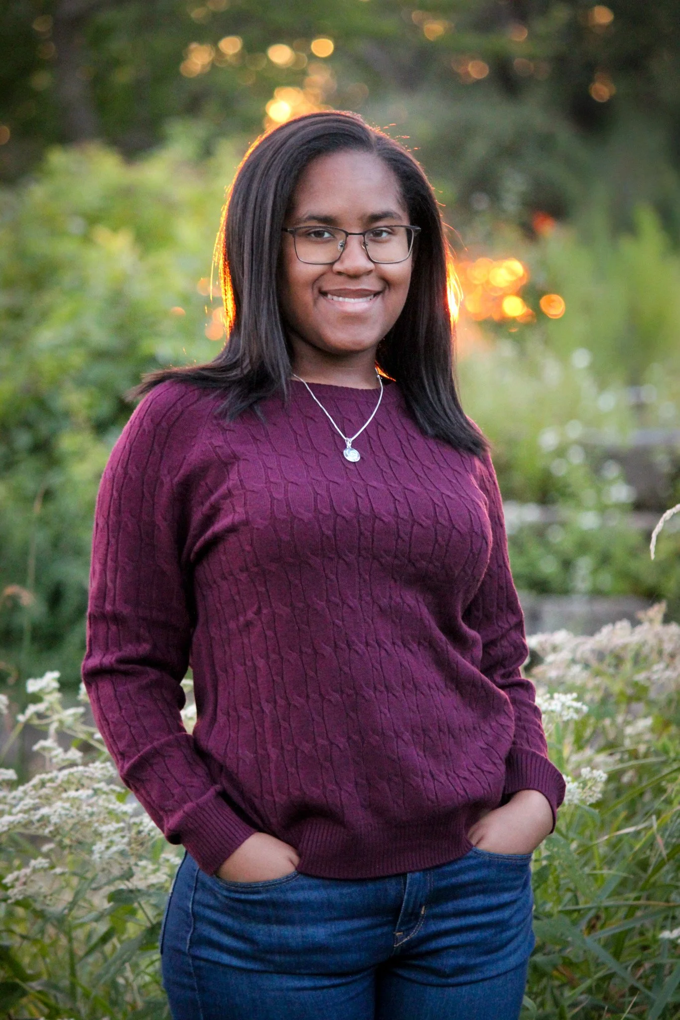 A young woman with glasses standing outdoors in a natural setting during sunset, smiling and wearing a maroon sweater and blue jeans.
