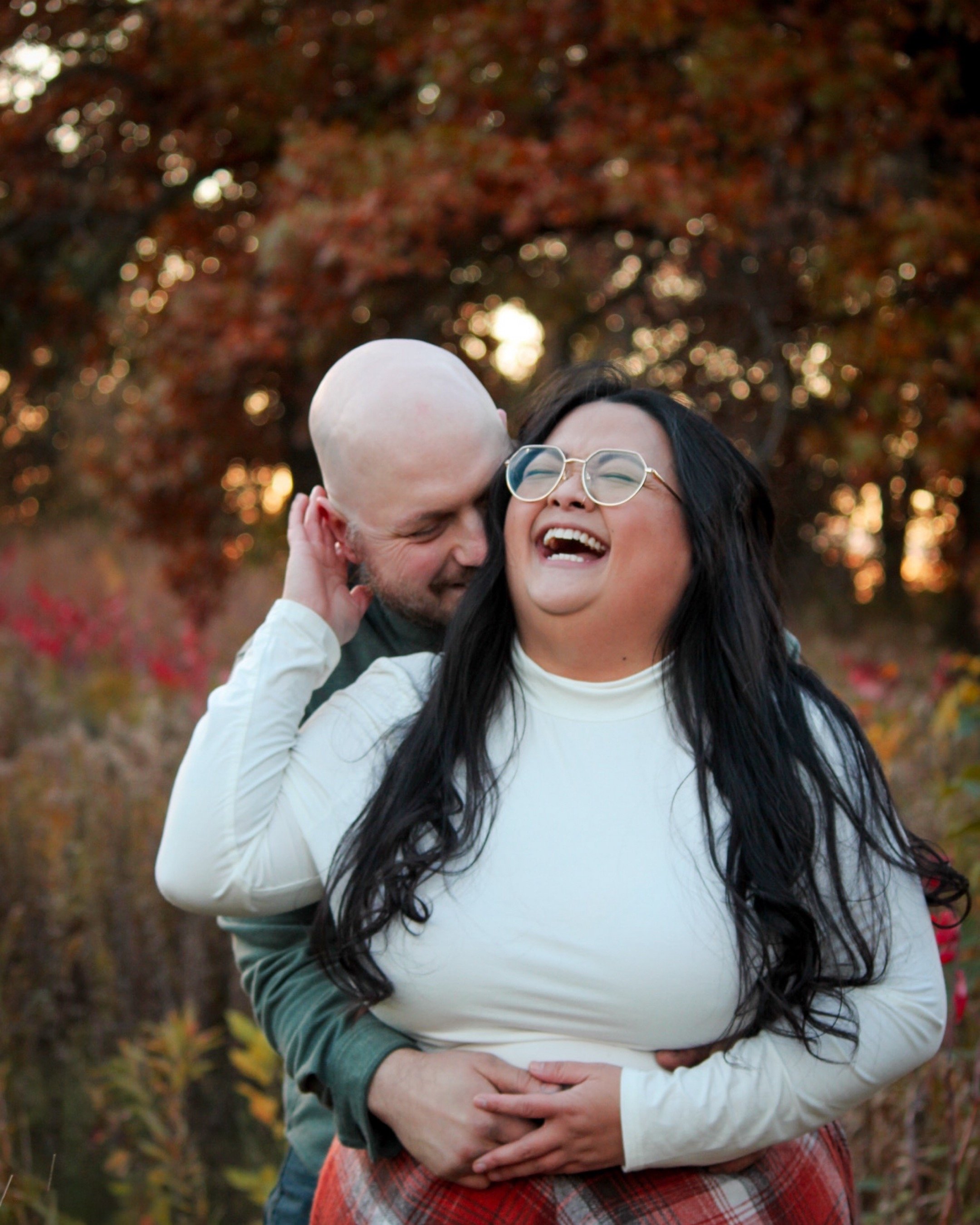 A couple laughing and embracing outdoors during autumn, with colorful fall trees in the background.