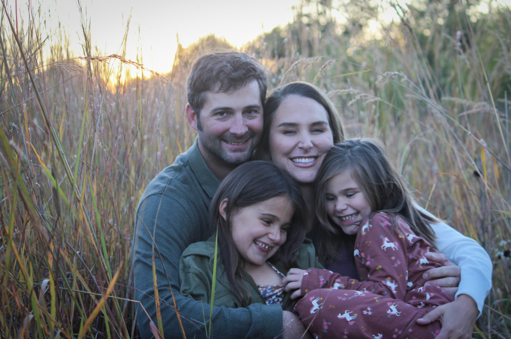 A family of four, consisting of a father, mother, and two young daughters, is sitting together in a field of tall grass during sunset, smiling and cuddling.