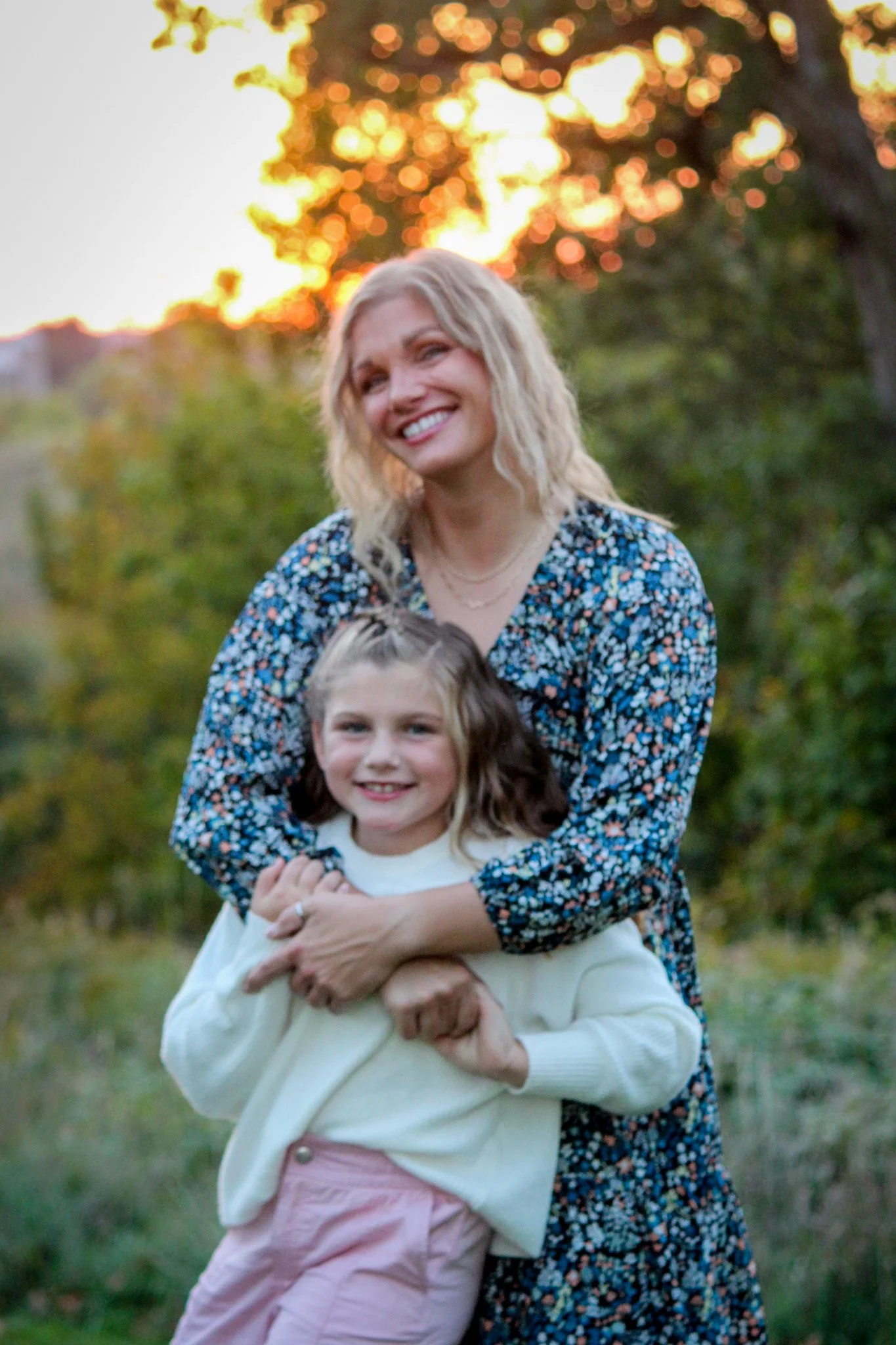 A smiling woman with blonde hair wearing a floral dress is hugging a young girl with brown hair in a white sweater and pink pants. They are outdoors during sunset with trees and a warm glow in the background.