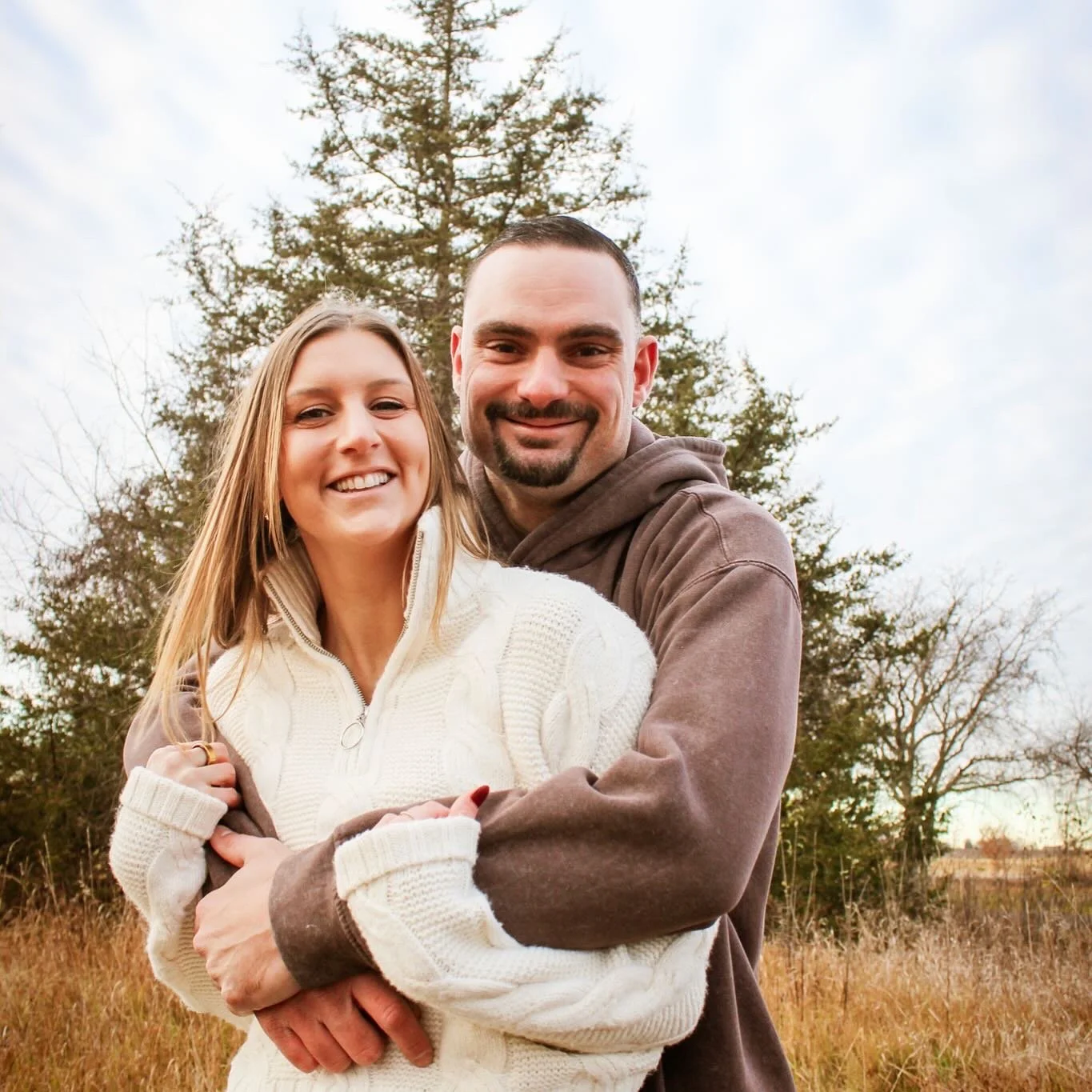 A smiling man and woman standing outdoors in a field with trees in the background, hugging each other.