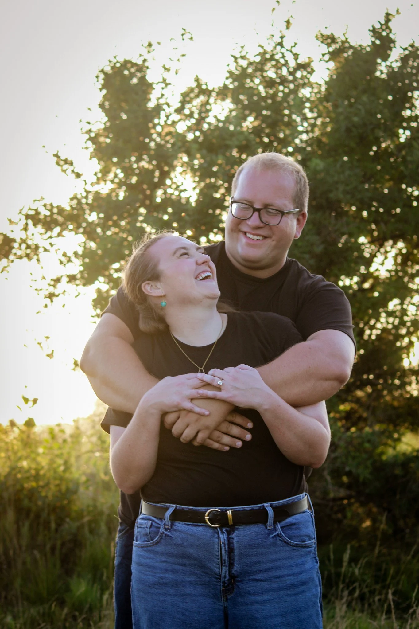 A happy couple outdoors, smiling and embracing each other, with trees and sunlight in the background.