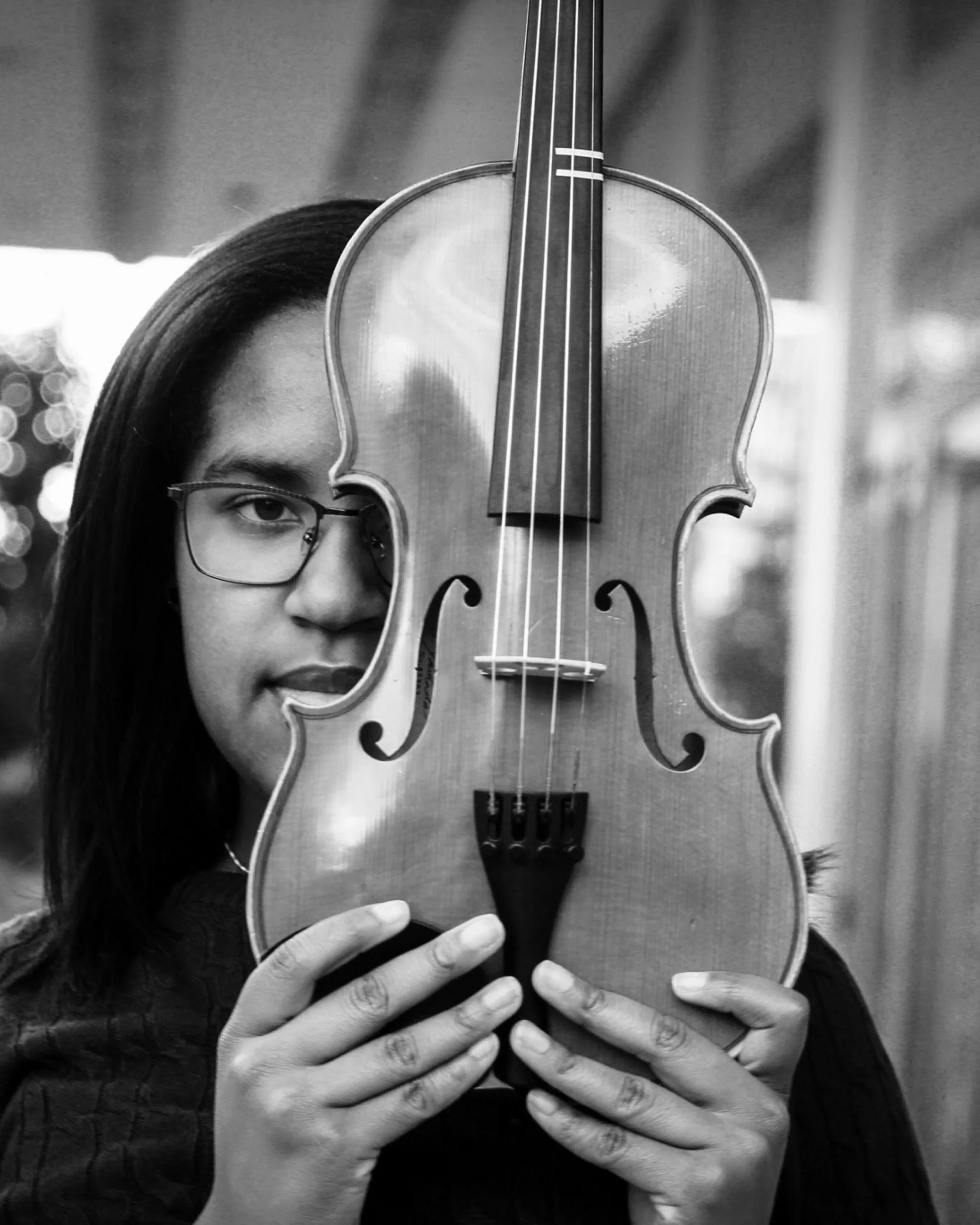 A woman with glasses holding a violin vertically in front of her face, with part of her face visible behind the instrument, in black and white.