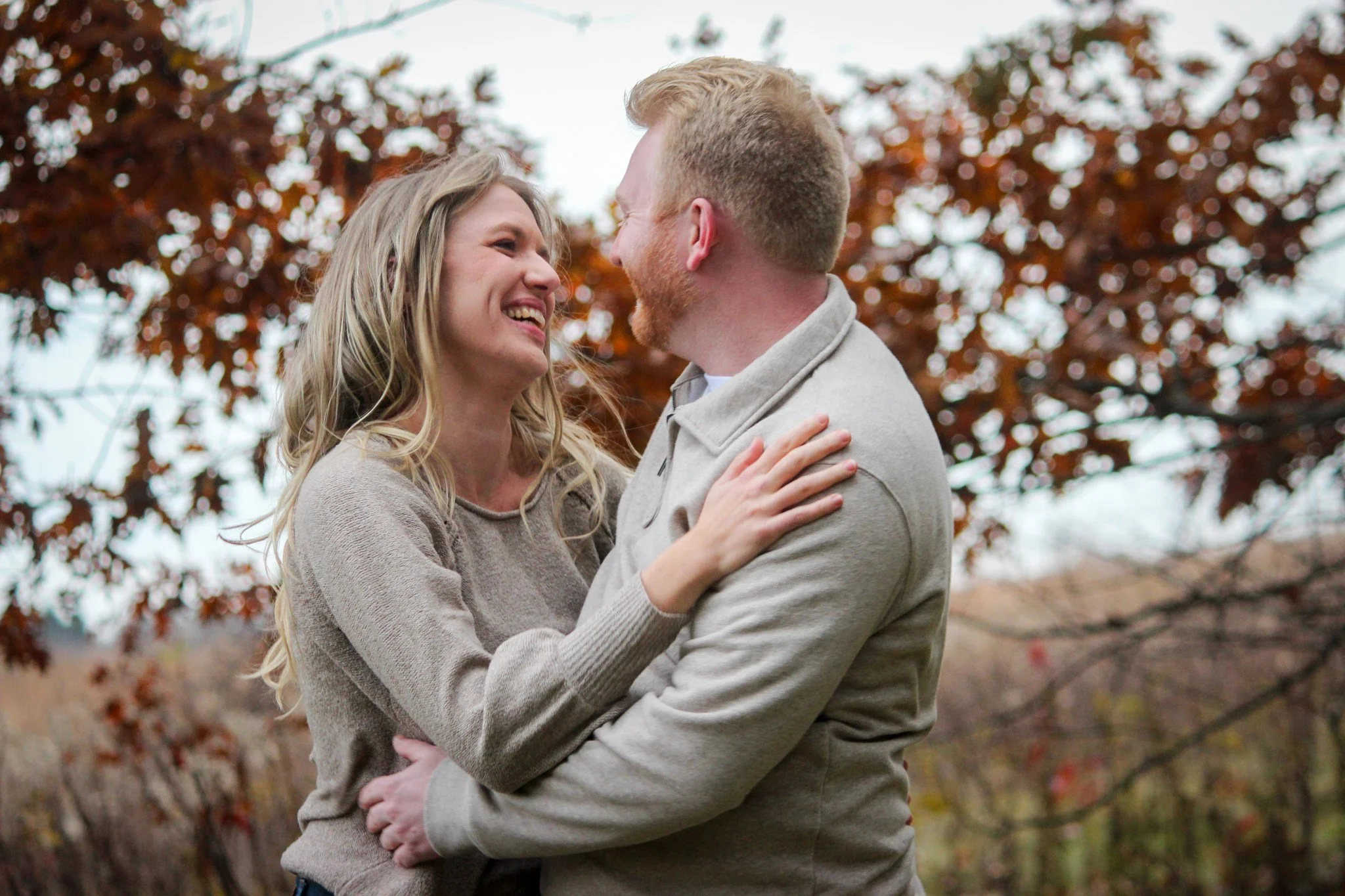A couple standing outdoors, smiling and looking at each other with a background of autumn leaves.