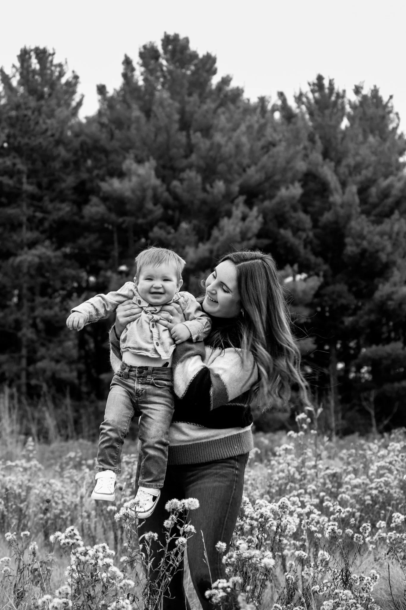 A woman holding a smiling child in a field of flowers with trees in the background, black and white photo.