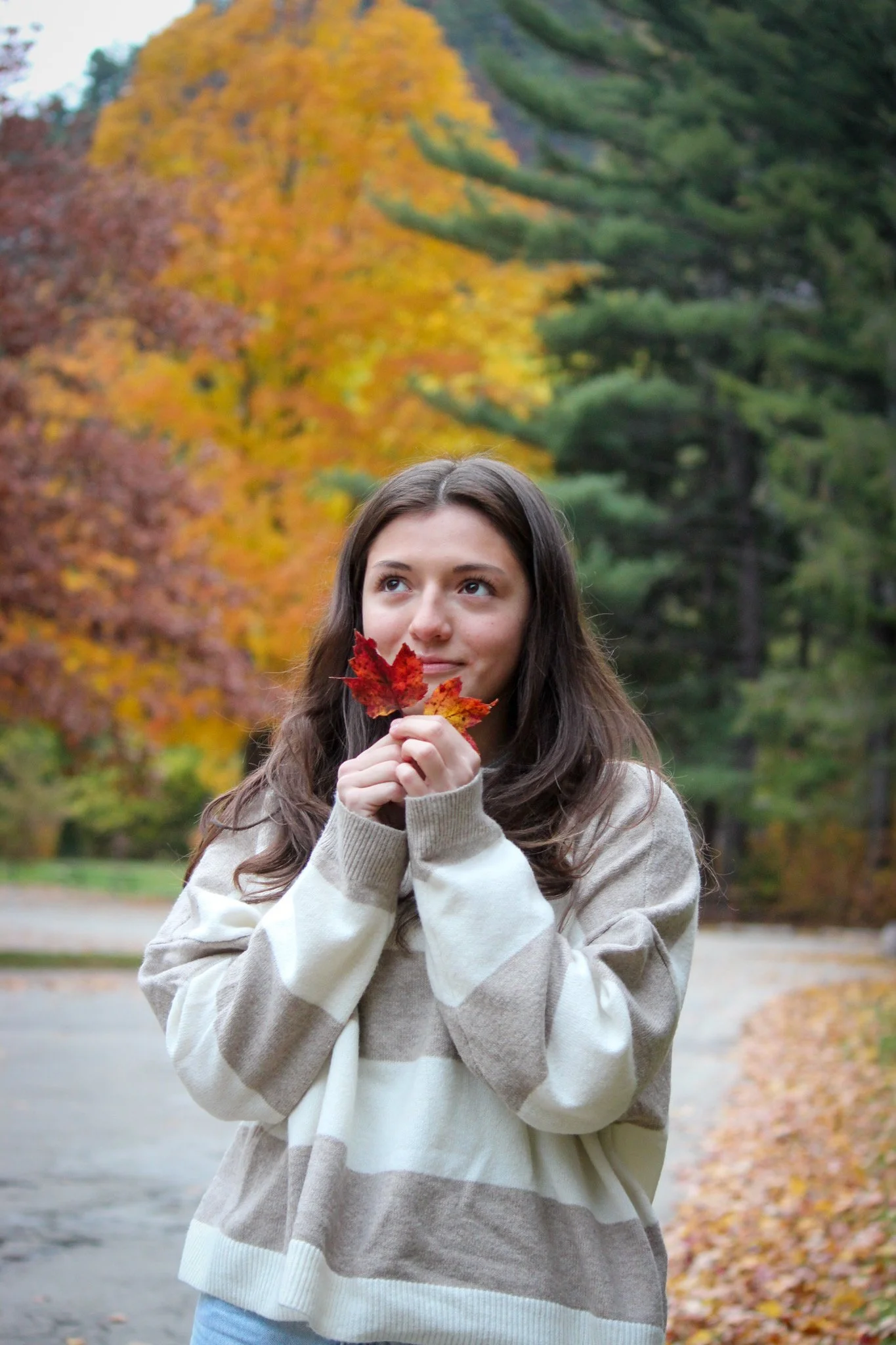 A young woman with long brown hair holding autumn leaves close to her face outdoors behind trees with fall foliage.