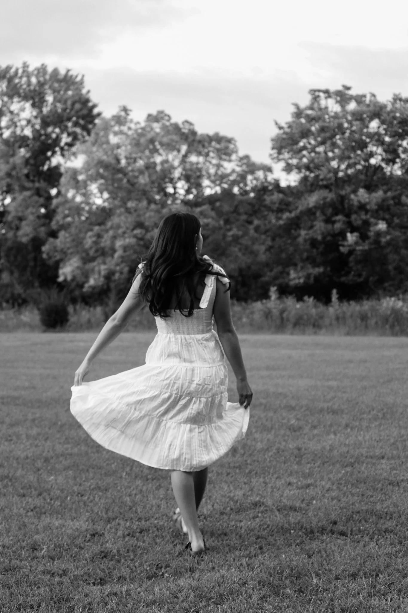 A woman wearing a white dress walking on grass in a park with trees in the background, captured in black and white.