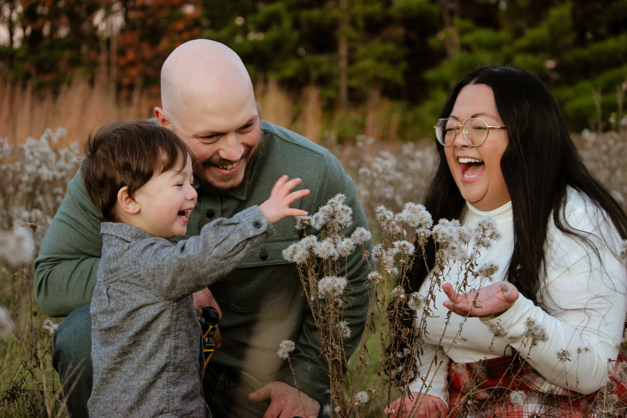 A family of three, a father, mother, and young son, laughing and enjoying time together in a field of wildflowers.