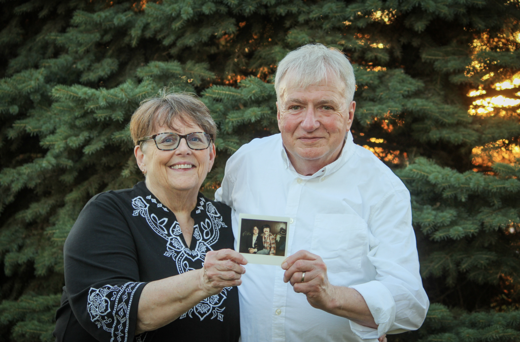 An elderly woman and man standing outdoors in front of a pine tree at sunset, smiling, holding a photograph of themselves together.