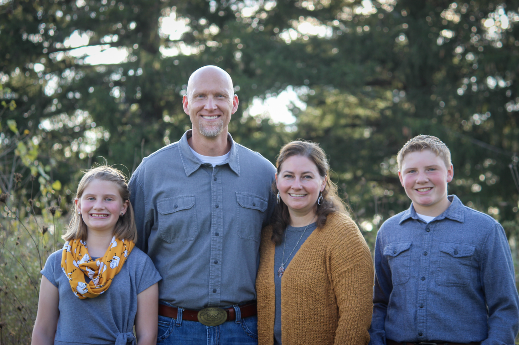A family of four standing outdoors in front of a wooded background. The father, mother, a girl, and a boy are smiling at the camera. The girl wears a gray top with a yellow scarf, the father has a gray button-up shirt, the mother has a mustard-colore