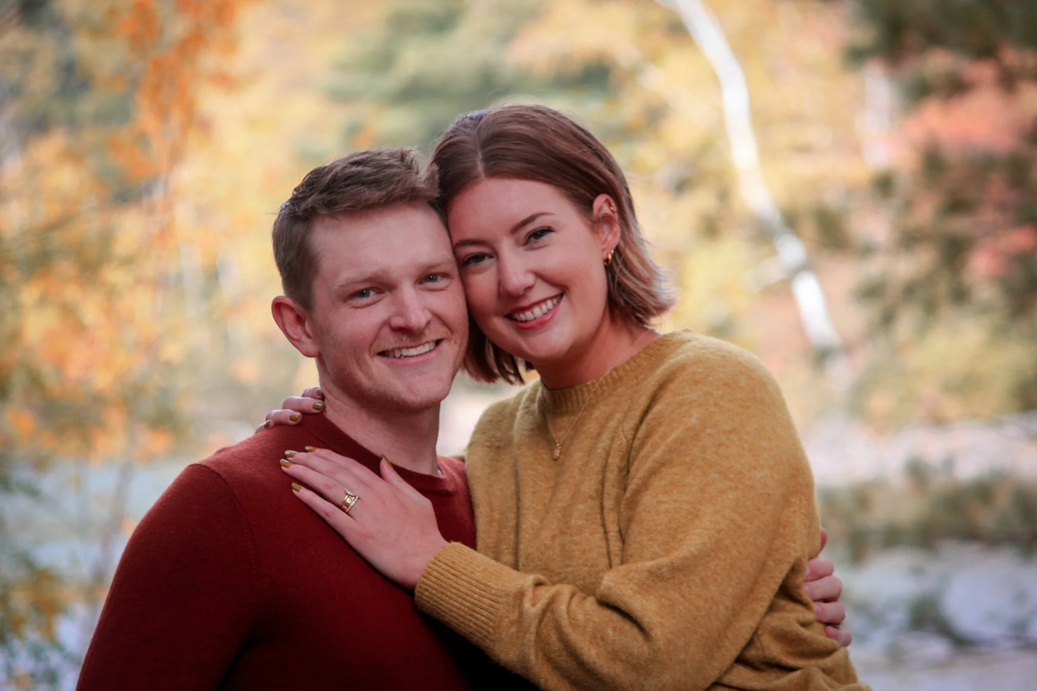 A couple smiling and embracing outdoors during autumn, surrounded by colorful fall foliage.