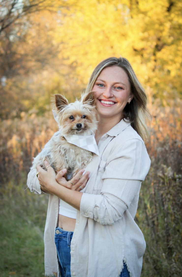 A woman with blonde hair smiling and holding a small Yorkshire Terrier dog outdoors during autumn, with yellow and orange trees in the background.