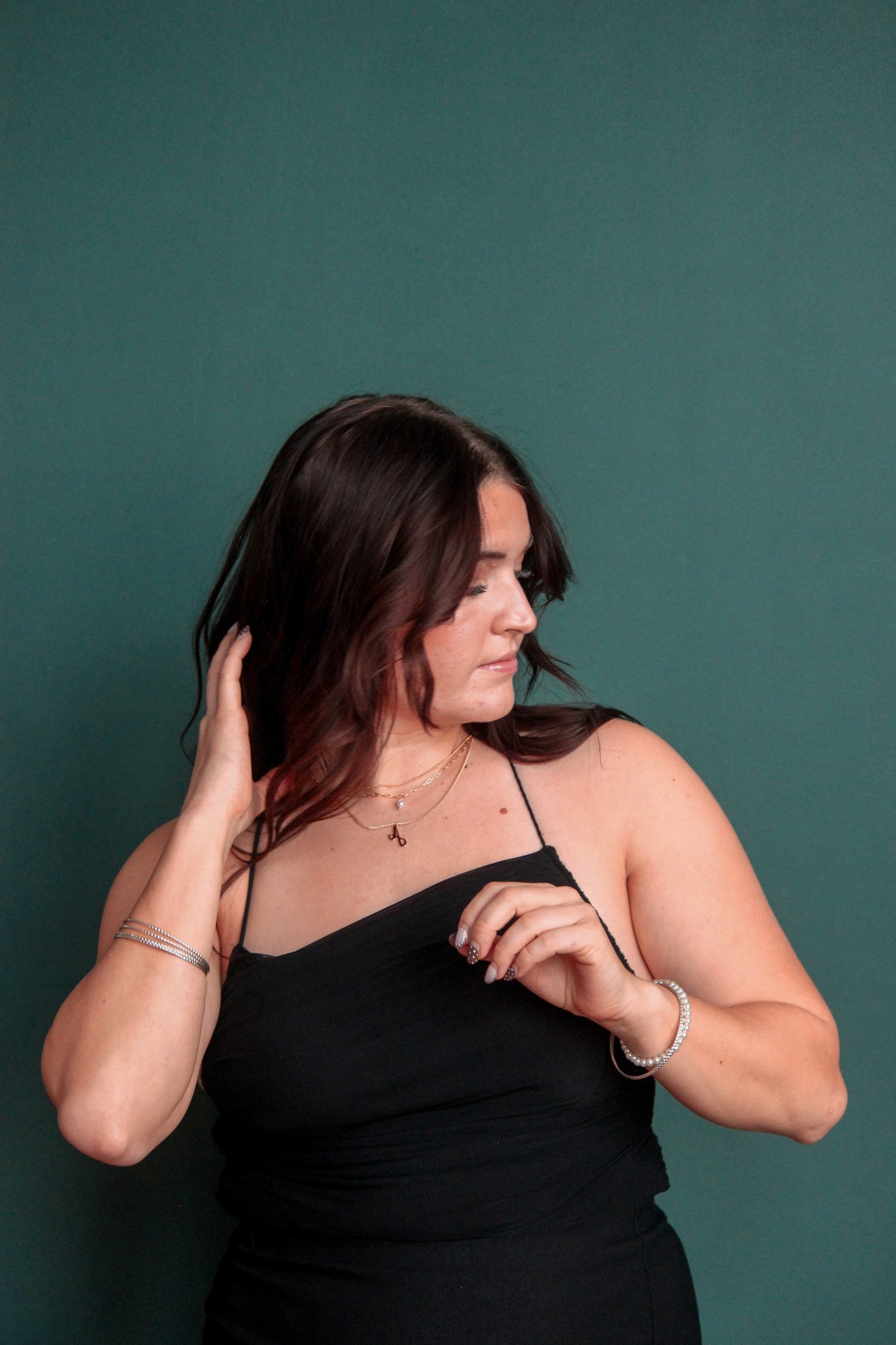A woman with dark brown hair in loose waves wearing a black spaghetti strap top, silver jewelry, and a neutral makeup look, standing against a green background, touching her hair with one hand and adjusting her top with the other.