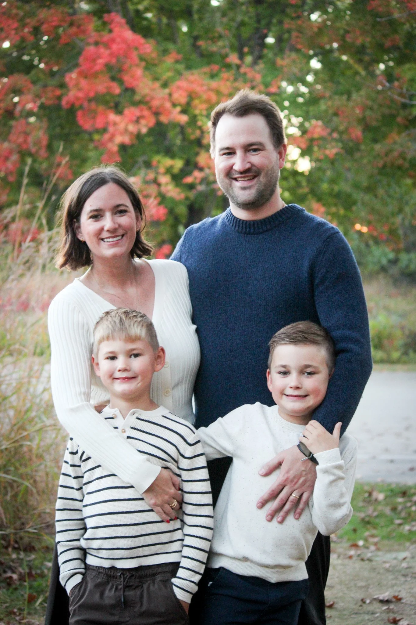 Family portrait outdoors during autumn, featuring two adults and two young boys smiling in front of colorful fall foliage.