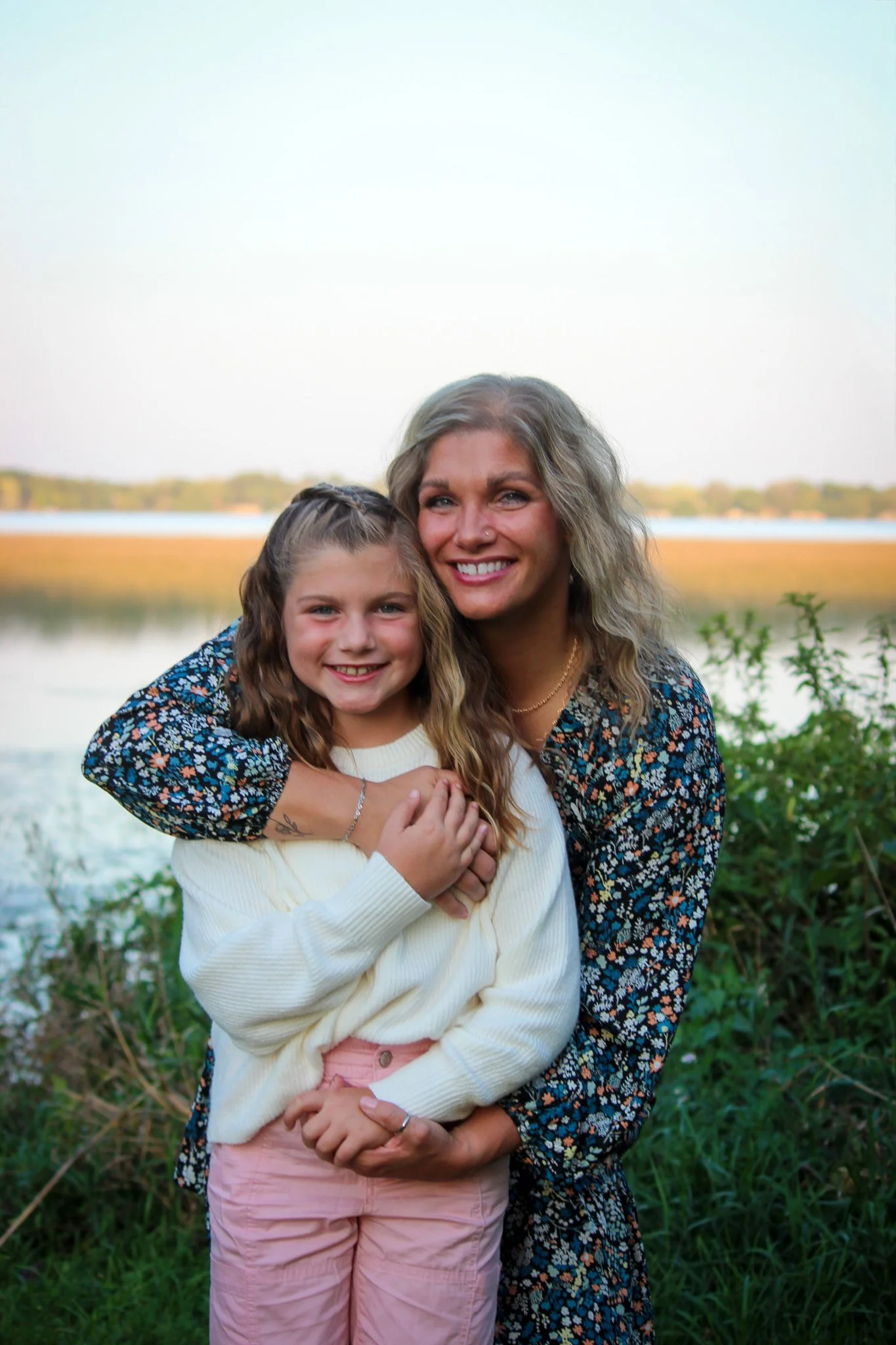 A woman and a girl smiling outdoors near a lake with greenery and a distant shoreline in the background.