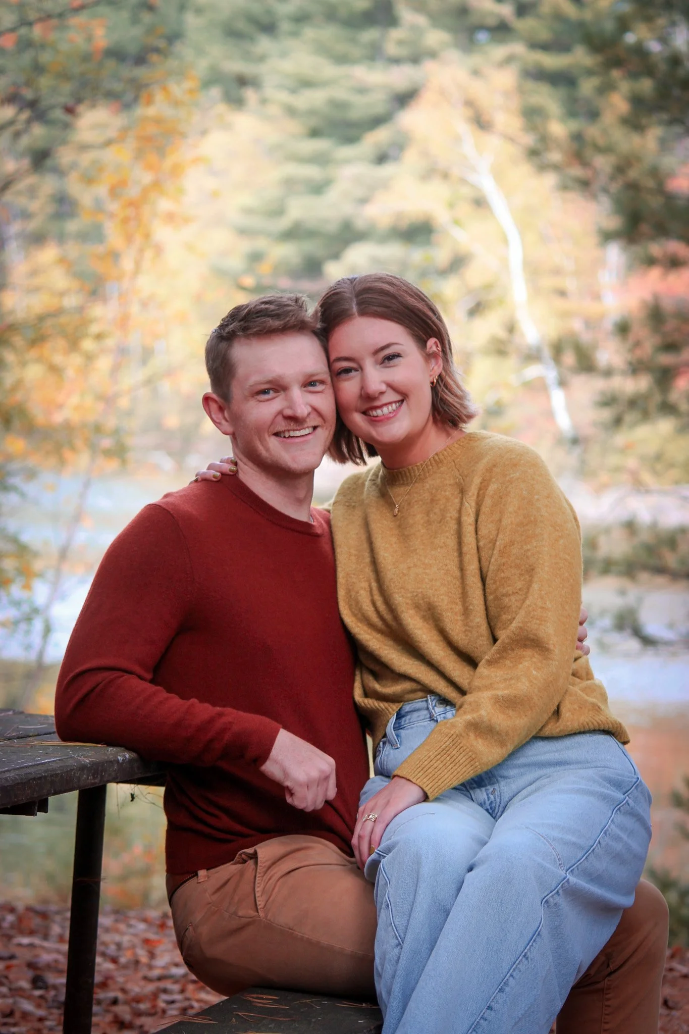 A couple sitting close together on a park bench outdoors during fall, with trees and autumn foliage in the background.