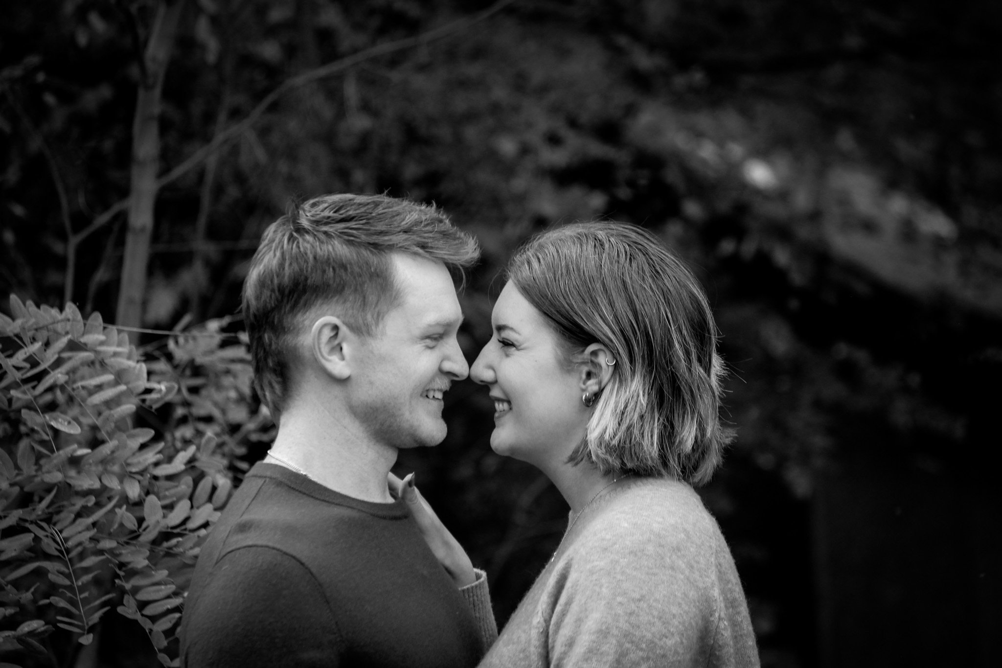 A black and white photo of a smiling young couple with their foreheads touching outdoors, surrounded by foliage.