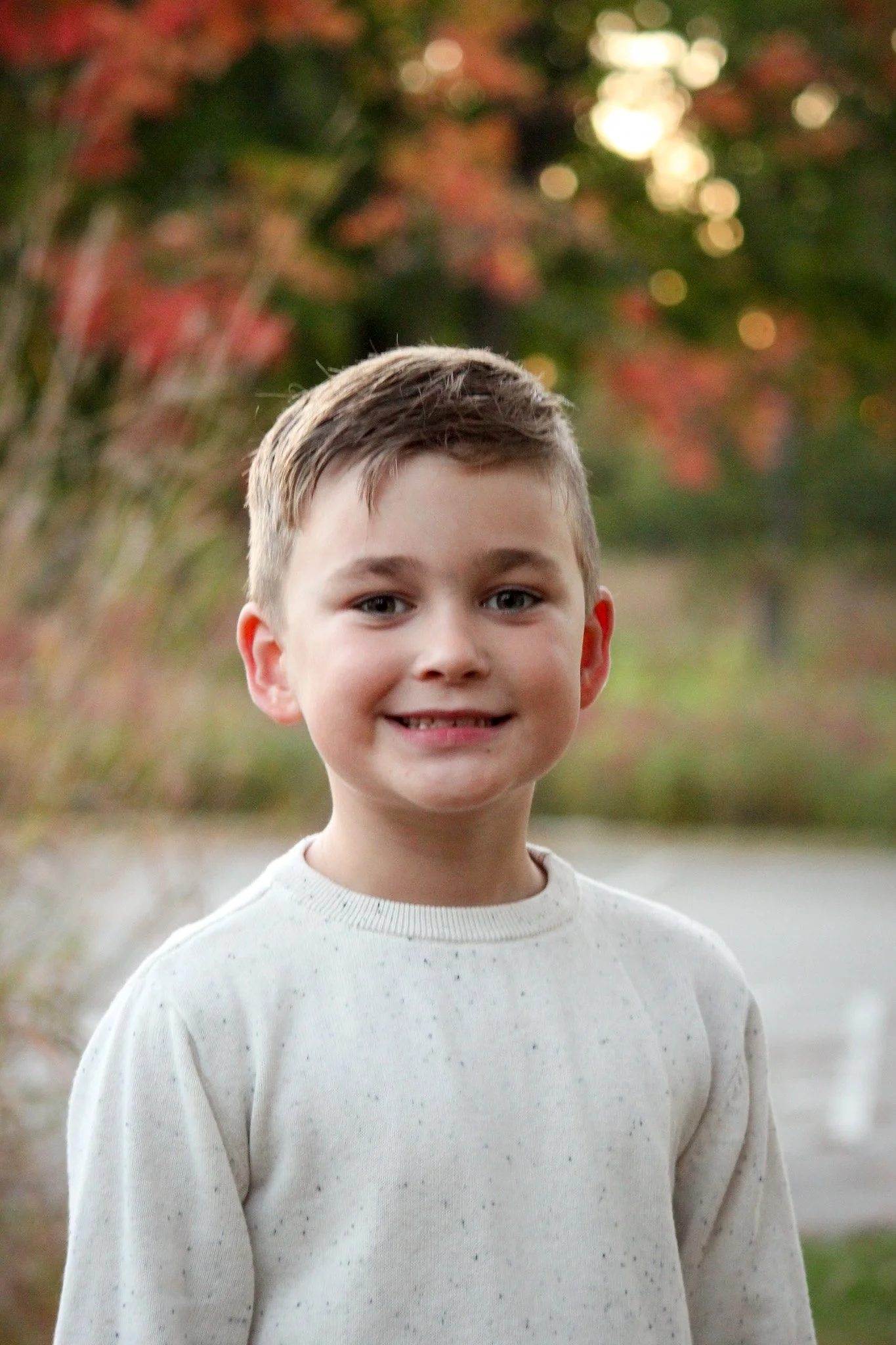 A young boy standing outdoors with trees in the background. He is smiling, wearing a cream-colored sweater, and has short brown hair.