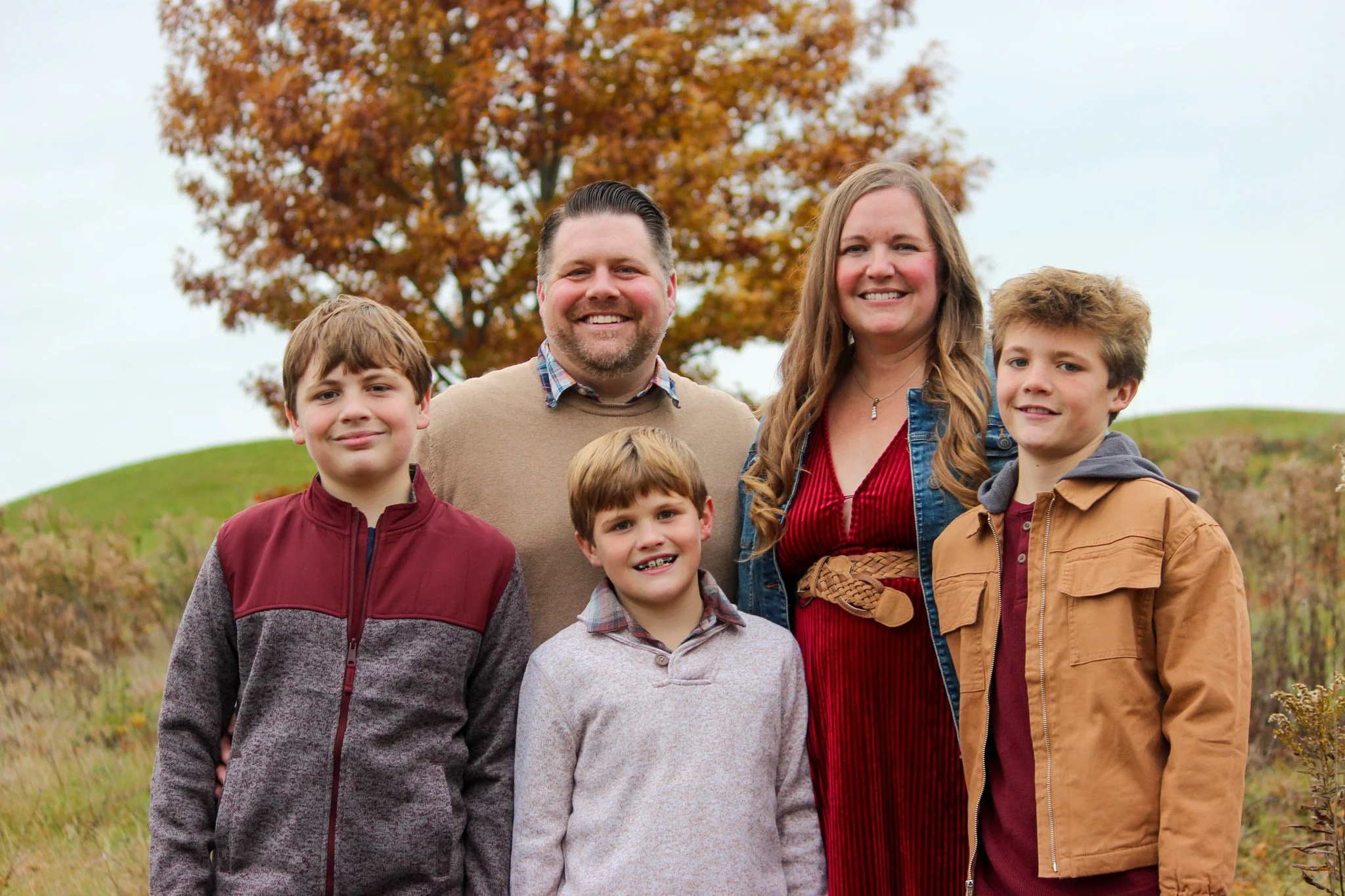 Family of five standing outside near a tree with orange leaves; two adults and three children smiling at the camera.
