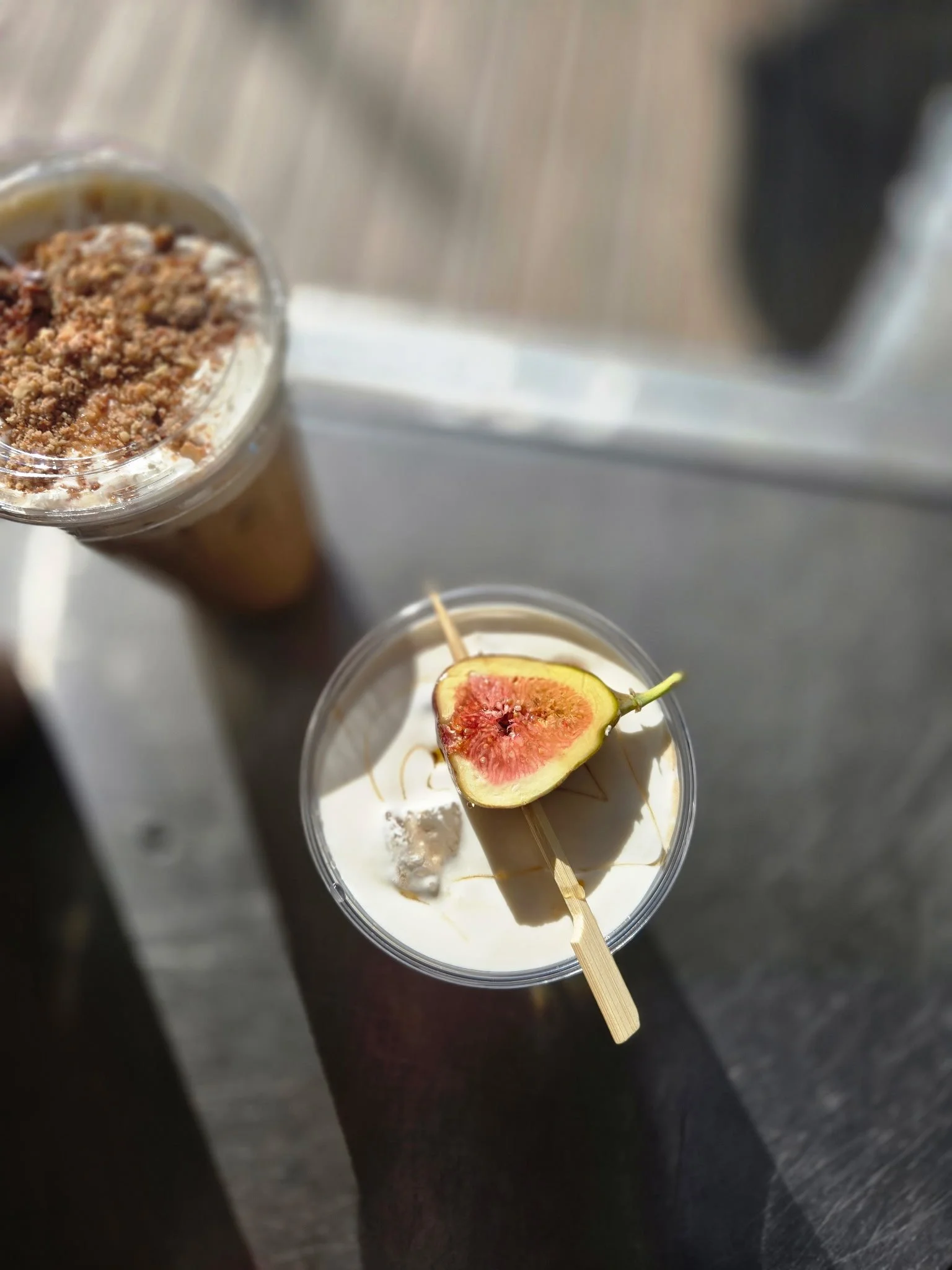 A clear plastic cup filled with a creamy dessert topped with a slice of fig and a wooden skewer, with another cup of similar dessert nearby under natural light.