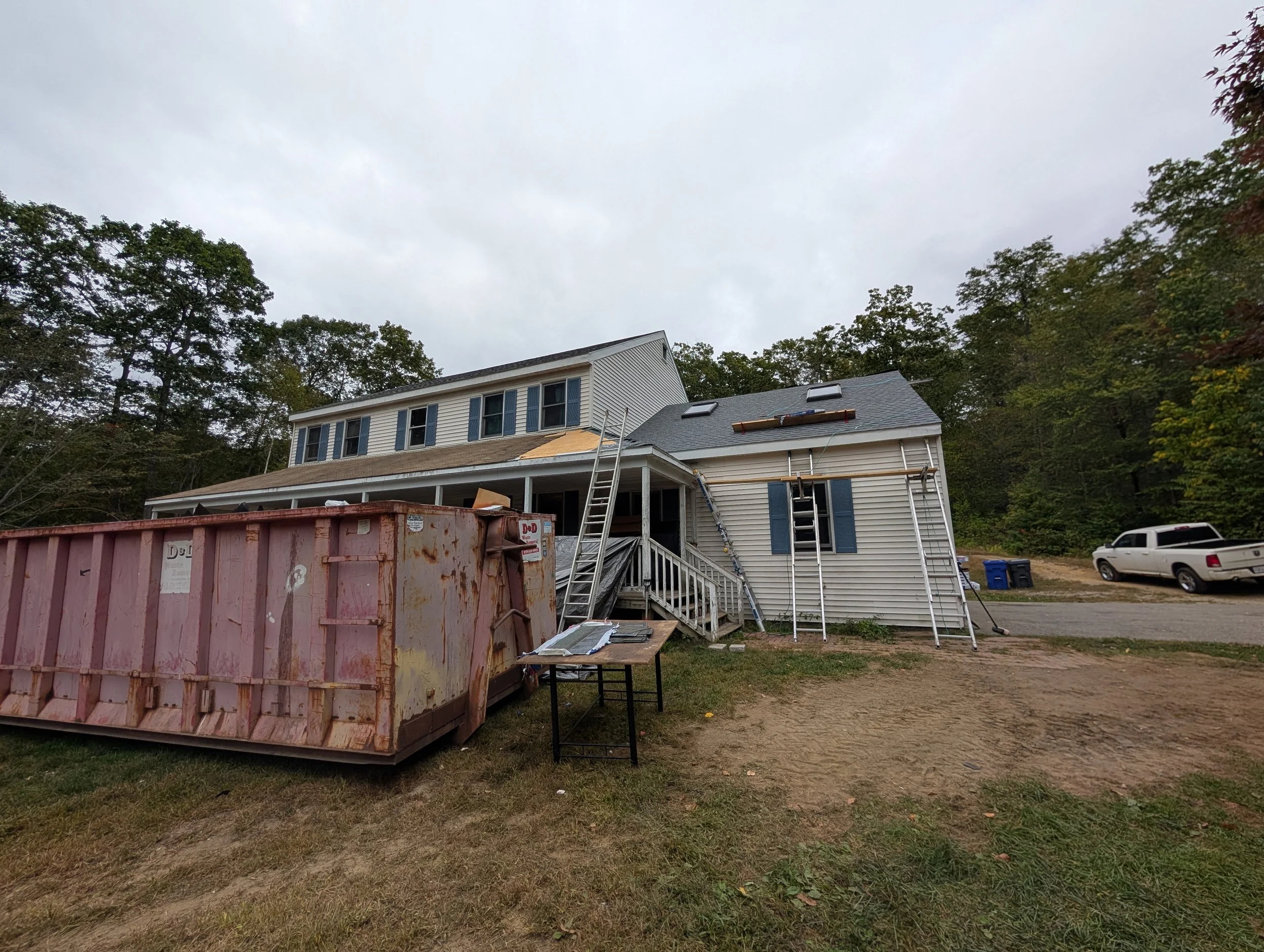 House under renovation with construction materials, ladders, and debris outside, a rusty dumpster in the foreground, and trees in the background.