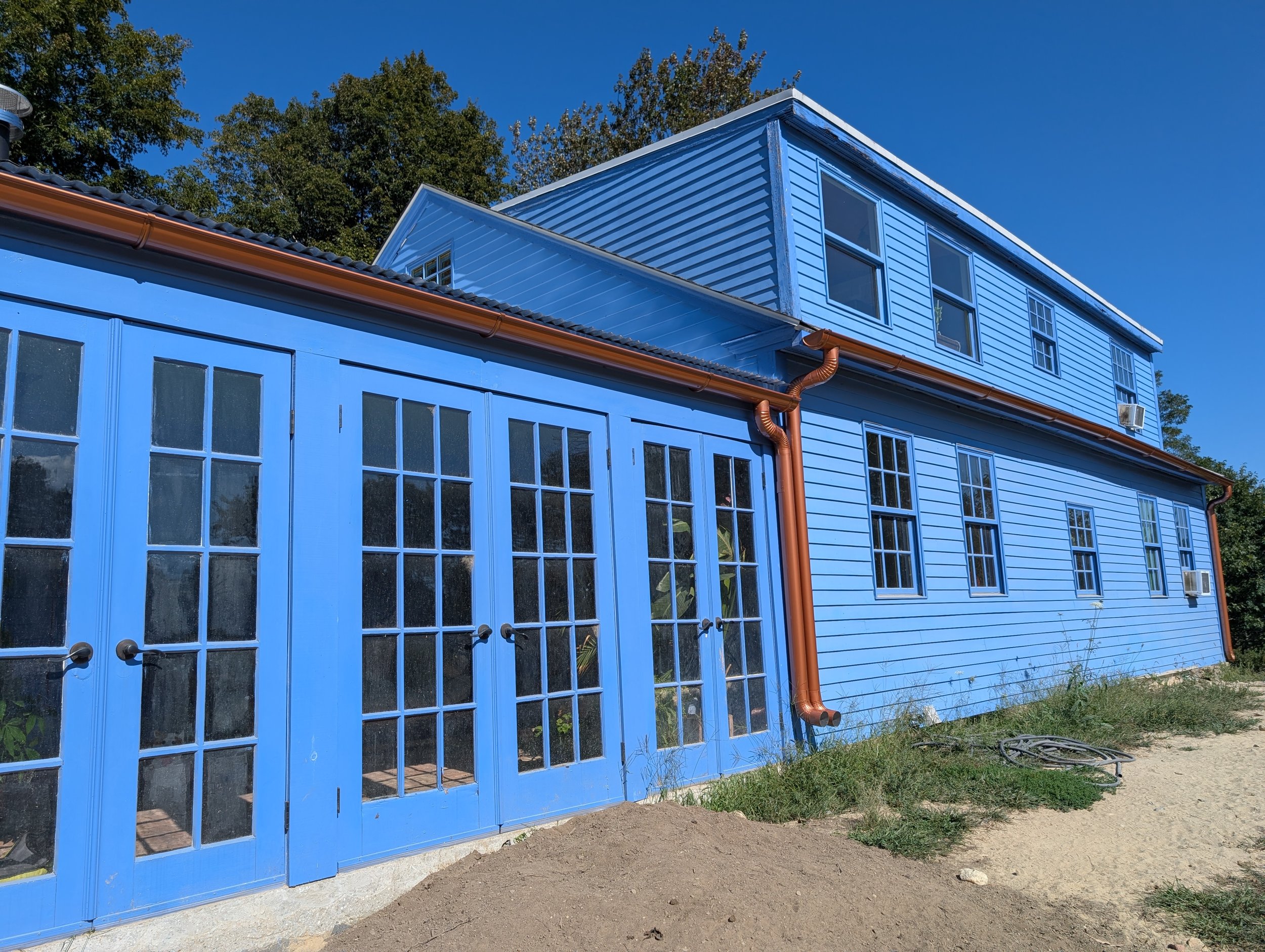 A two-story house painted in bright blue with multiple windows and a glass-paneled blue door. There are copper-colored gutters and downspouts, and an air conditioning unit is visible on the second floor. The house is set against a clear blue sky with trees in the background, and the ground is mostly dirt with some patches of grass.