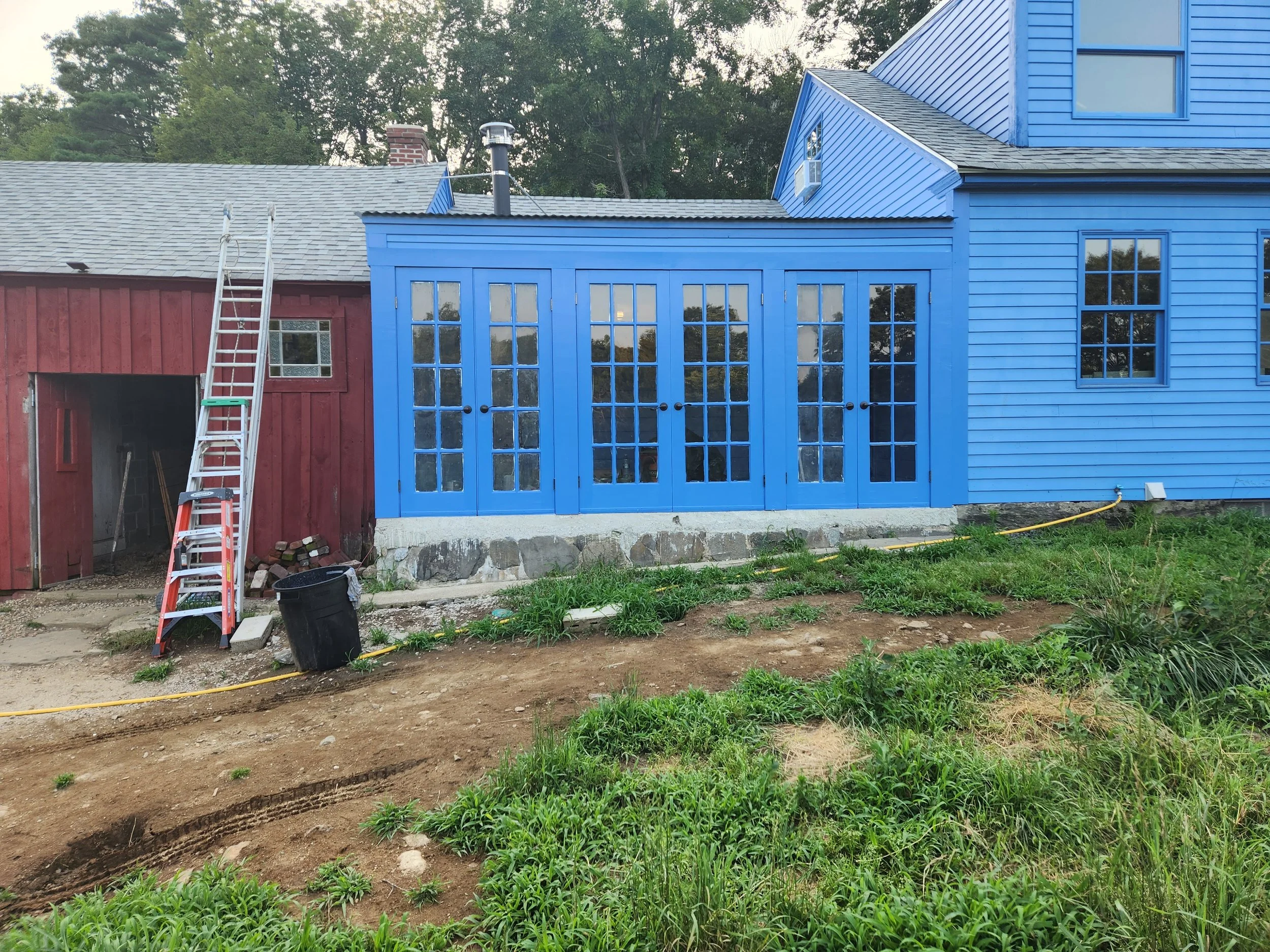 A house with blue siding and a sunroom with glass doors, next to an older red barn-like structure with a ladder leaning against it, on a grassy yard under an overcast sky.