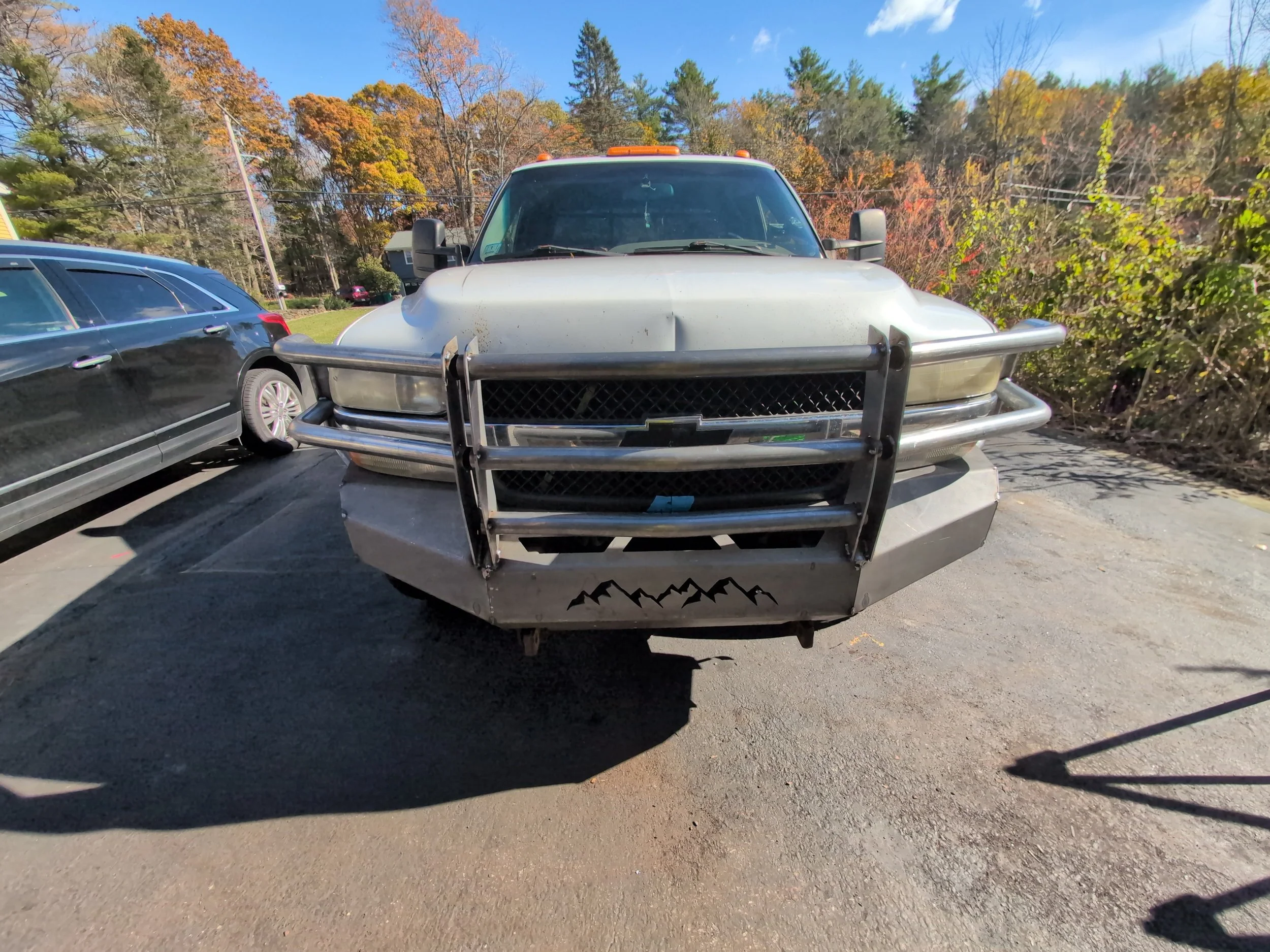 Front view of a white Chevrolet pickup truck with a large metal bumper and guard, parked next to a black vehicle, with trees showing fall foliage in the background.