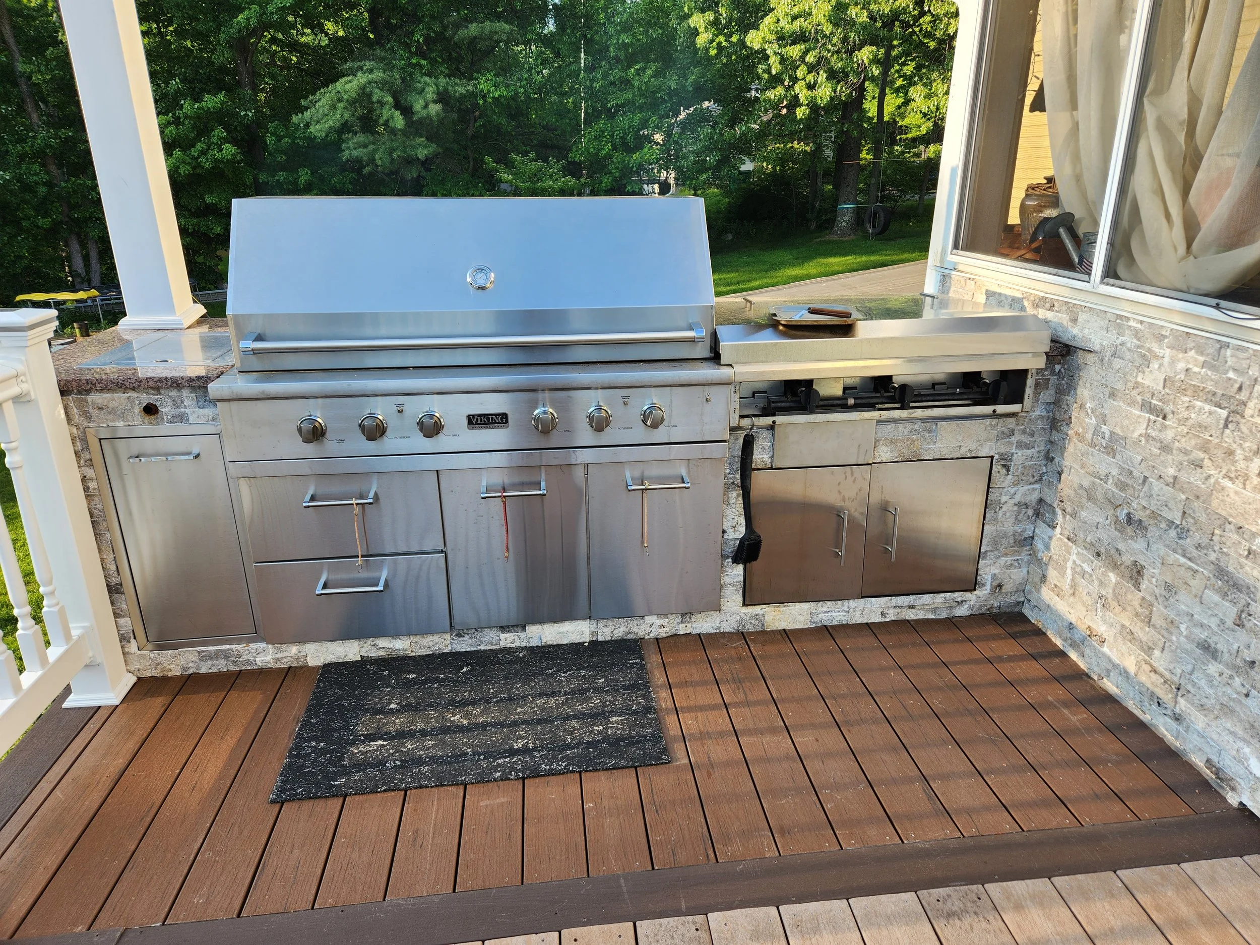 Outdoor kitchen with a stainless steel Viking grill, a side burner, and storage cabinets, situated on a wooden deck with a black mat, with trees and grass visible in the background.