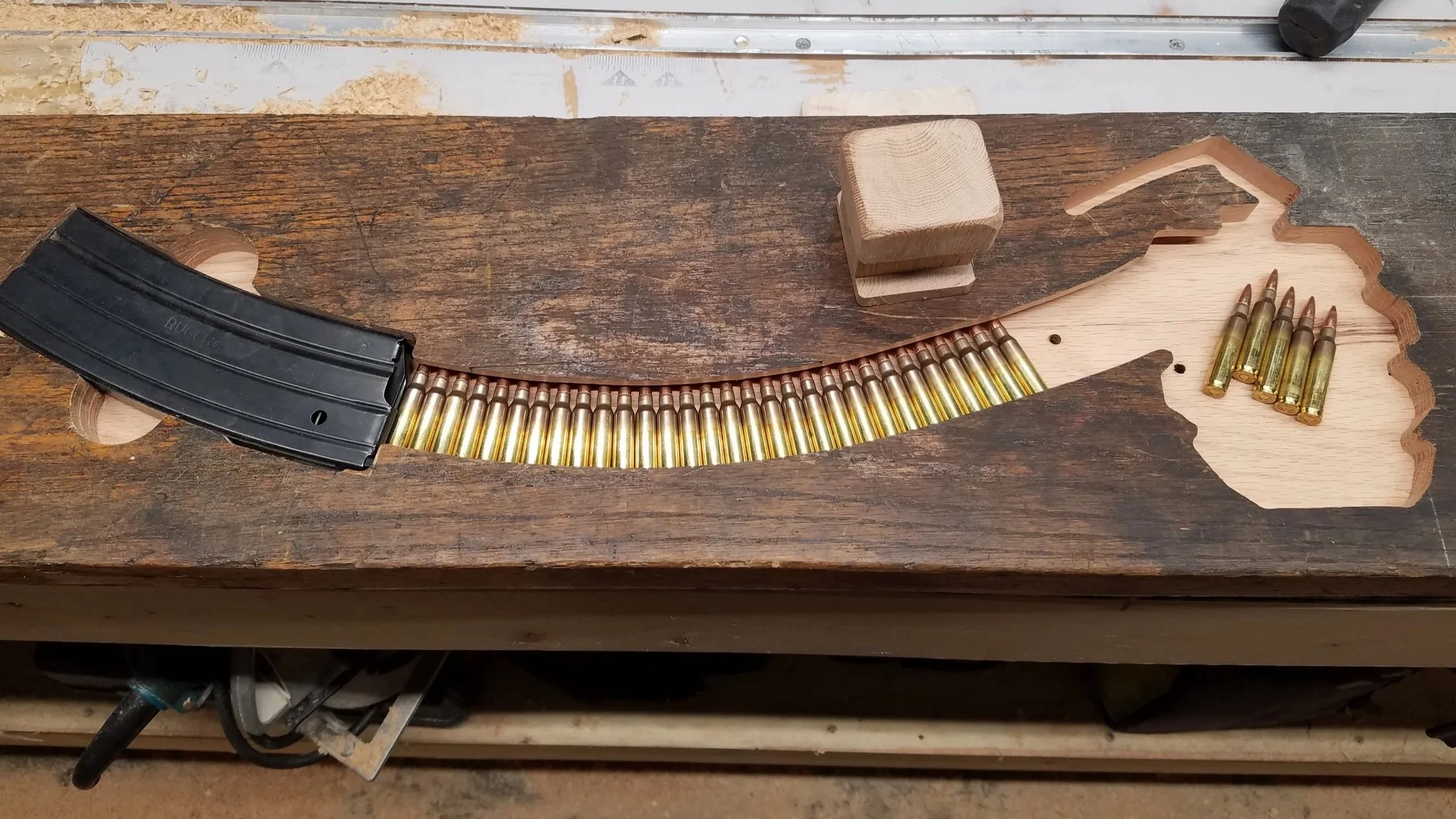 Partially completed wooden map of the state of Wisconsin with shot shell casings arranged along its outline, a black magazine, and a small block of wood on a workbench.