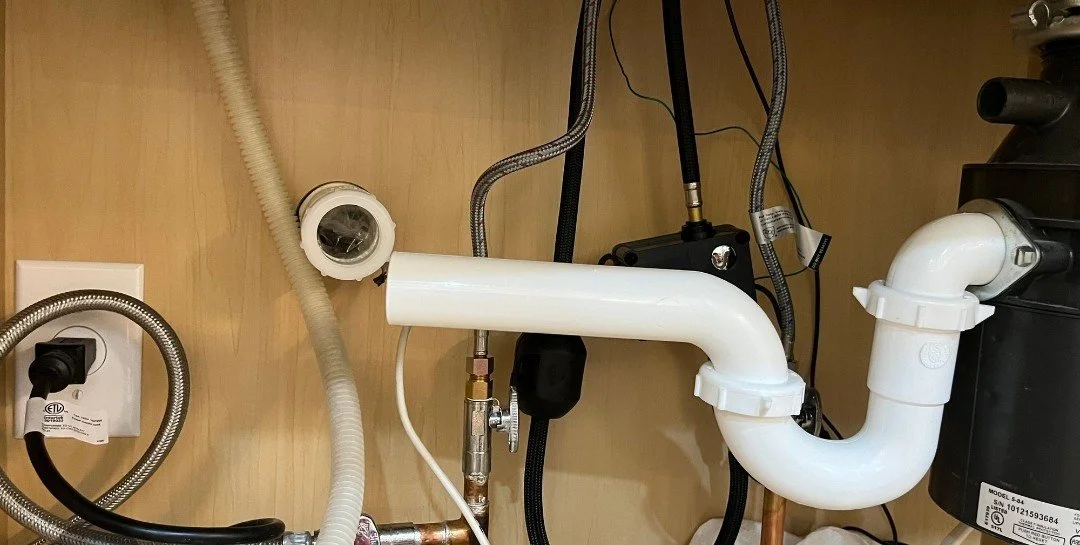 View of plumbing pipes, hoses, and electrical components under a sink or appliance, with white PVC pipes, metal hoses, and black plastic parts.
