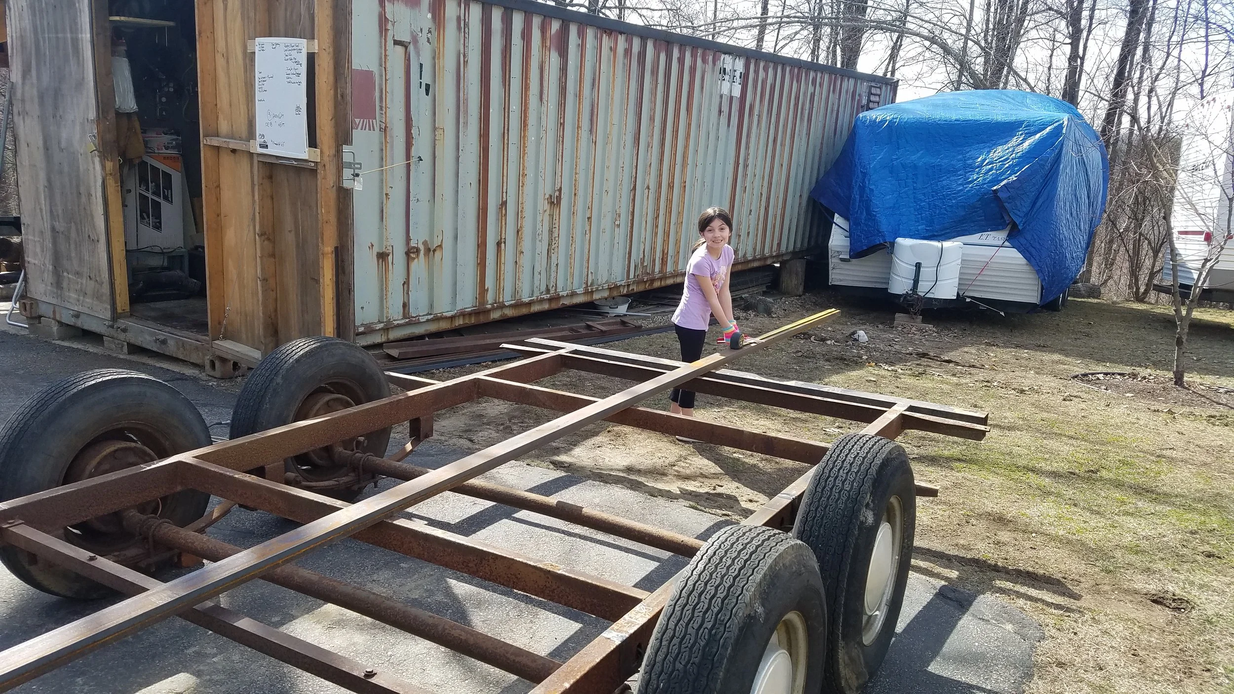 A young girl smiling and holding a measuring tape is standing on a metal frame with wheels in an outdoor yard. There are two large containers or trailers in the background, one rusted and the other covered with a blue tarp, with bare trees surrounding the area.