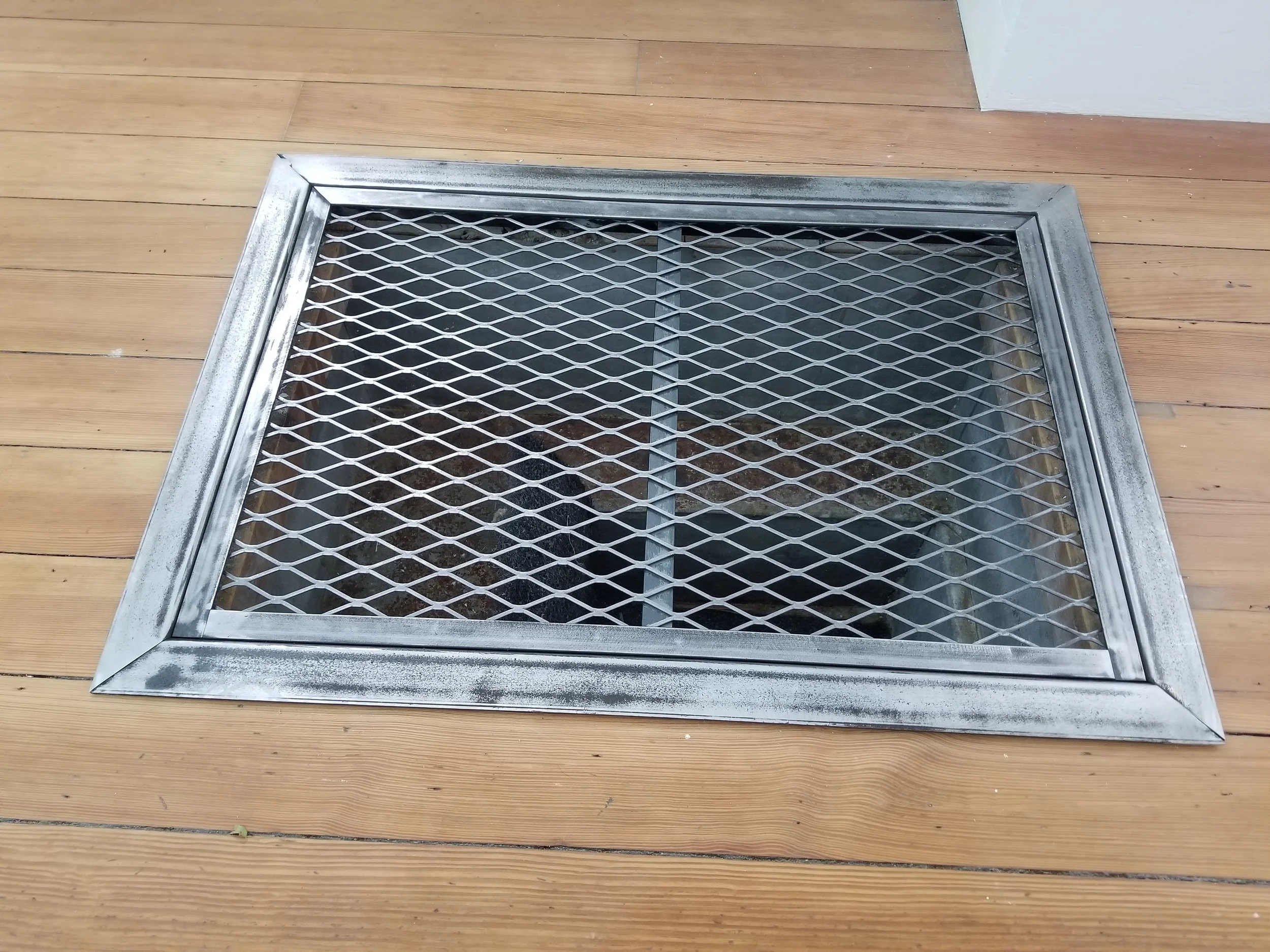 A metal vent cover on a wooden floor, with a metal grid over a vent opening underneath.