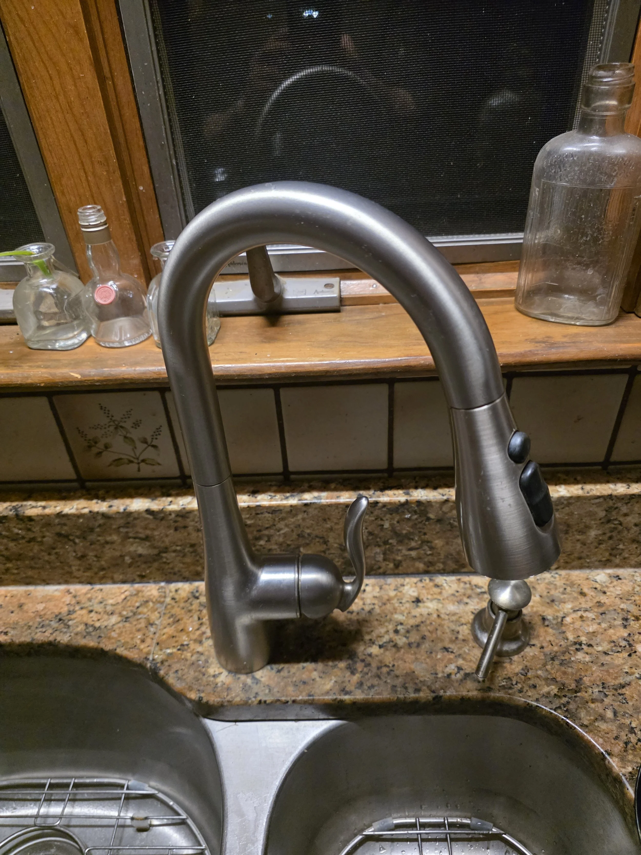 Kitchen sink with a modern stainless steel faucet, and countertop with several empty glass bottles, in front of a window with a screen.