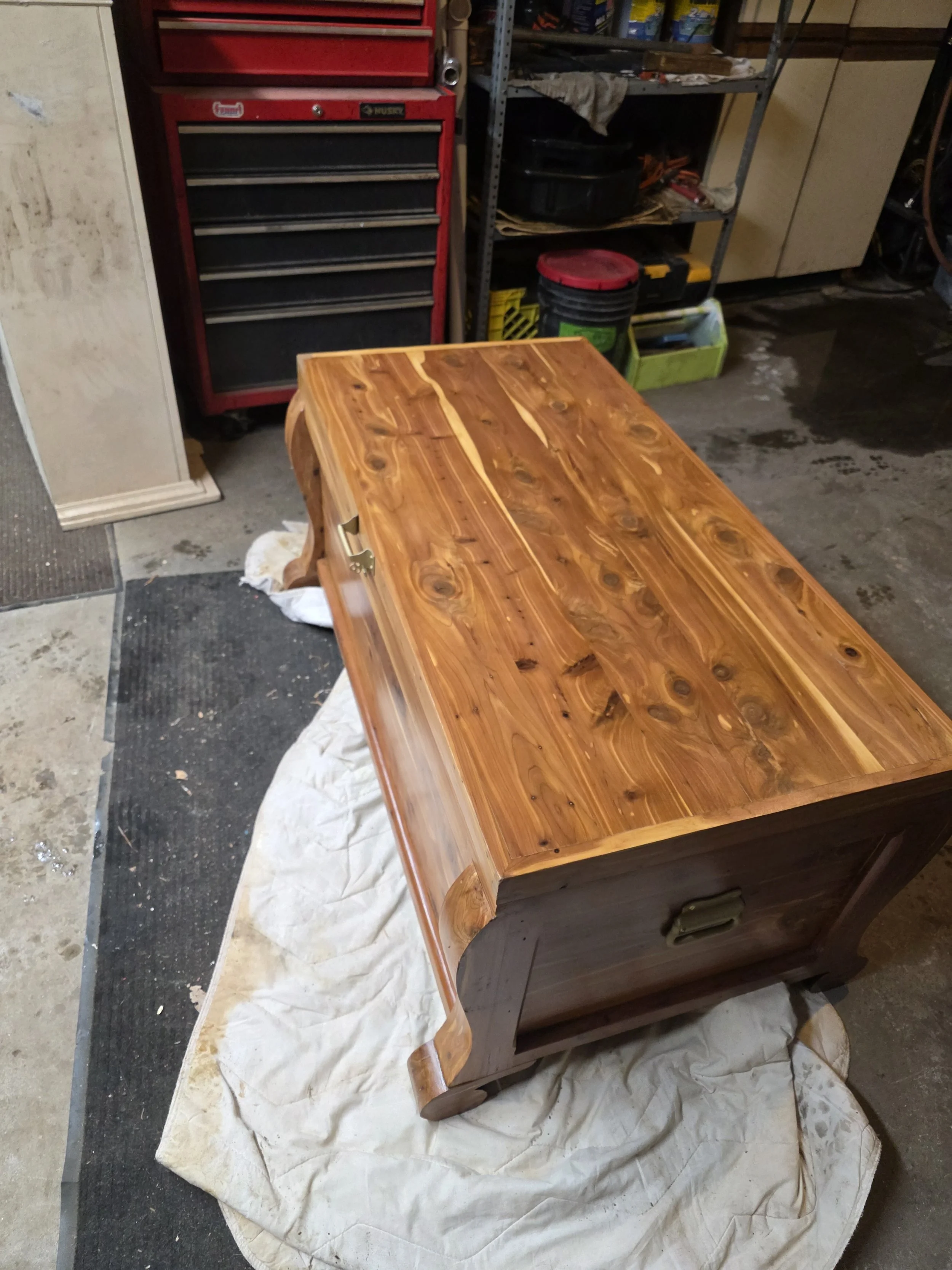 A wooden chest or bench with a polished finish in a workshop. It is placed on a white cloth on the floor, with tools and storage shelves visible in the background.