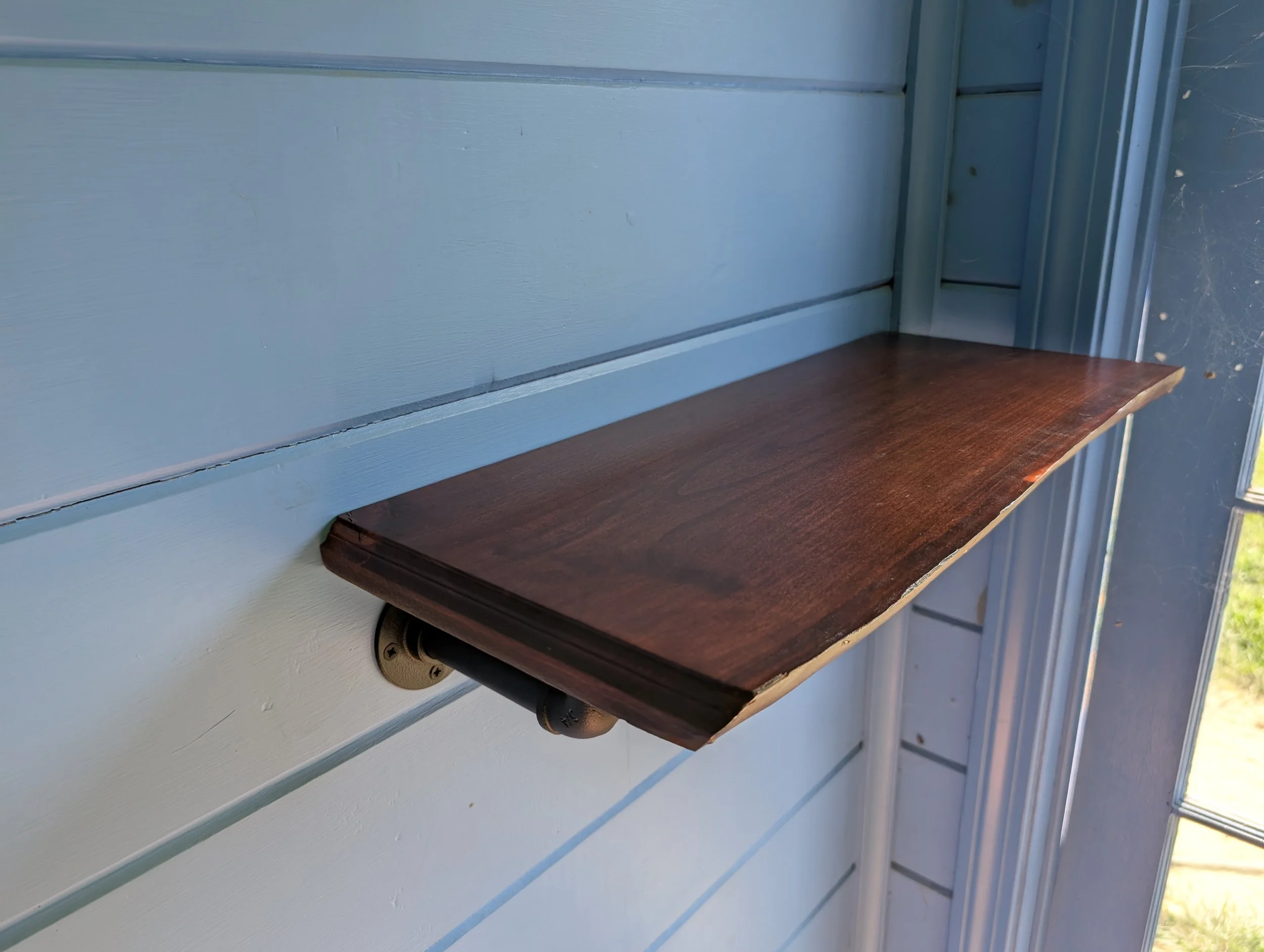 A wooden mailbox shelf attached to a light blue house wall with horizontal siding.