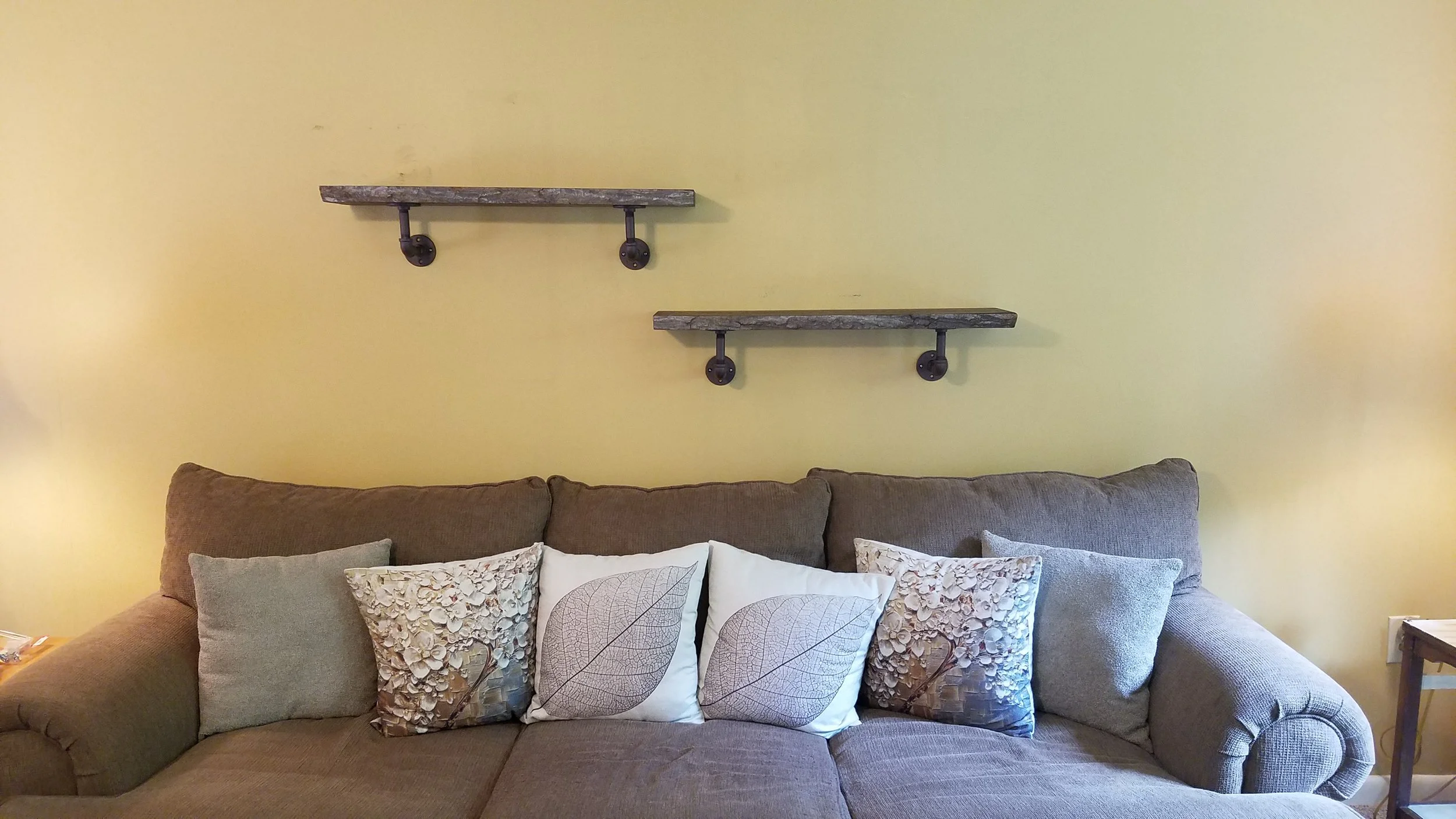 Living room with a brown sofa, decorative pillows, and two rustic wooden shelves mounted on a yellow wall.