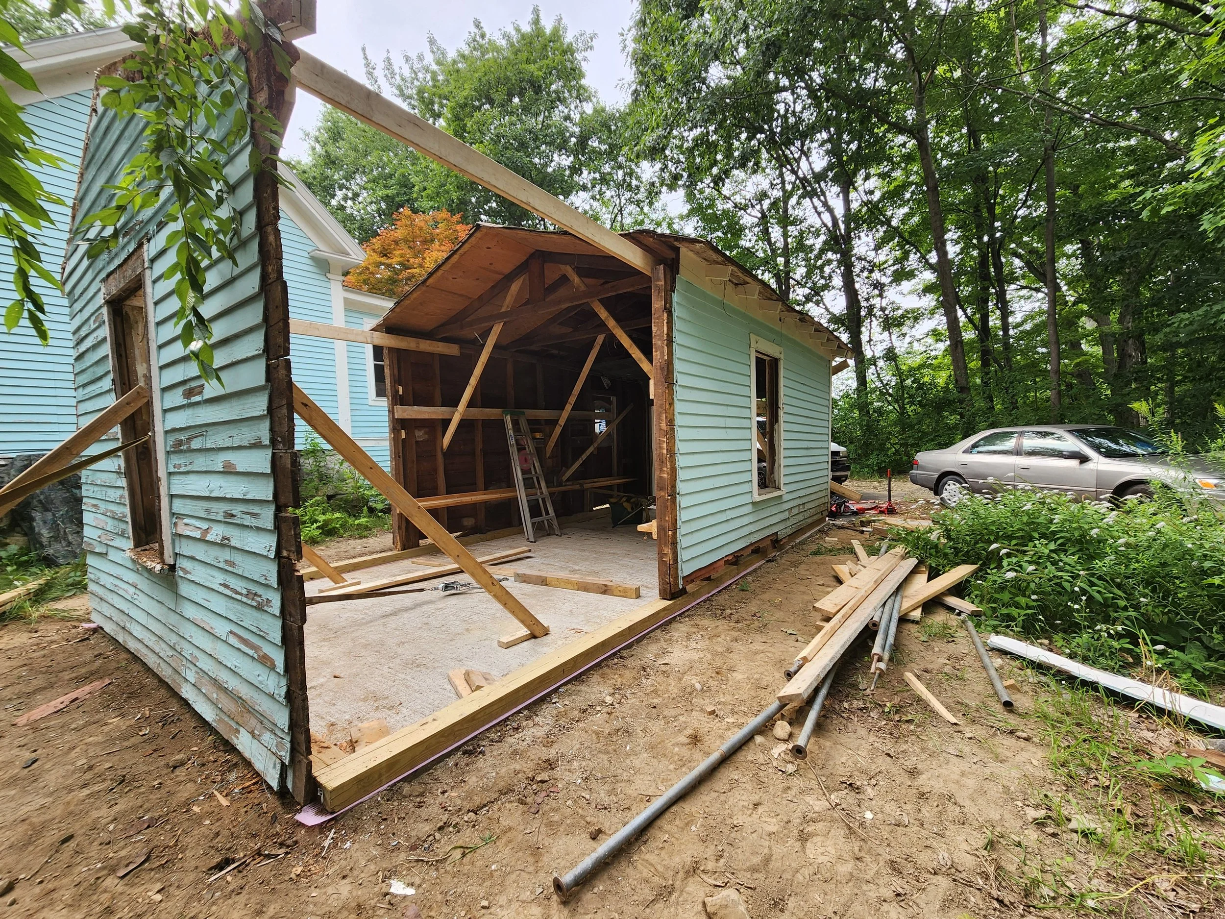 A house under renovation with siding being replaced, showing a partially demolished exterior wall and construction materials on the ground, surrounded by trees and parked cars.