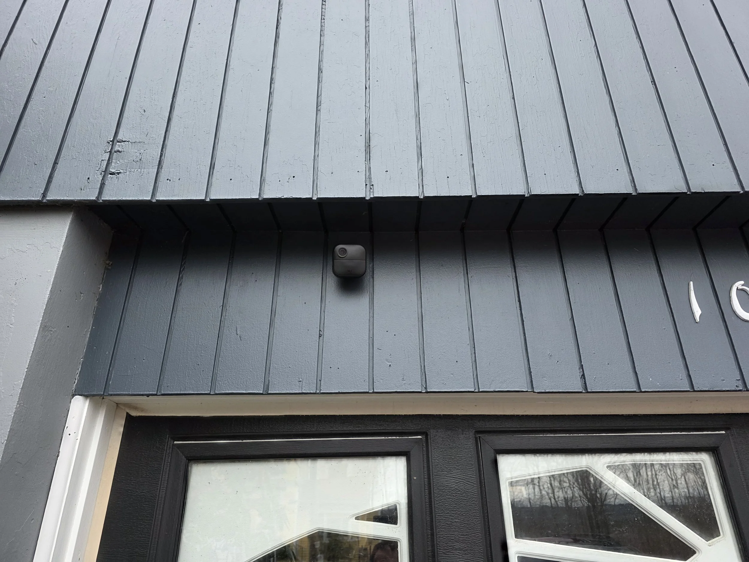 Close-up of a gray wooden exterior wall with siding, a small security camera, and part of a window with a reflection of trees.