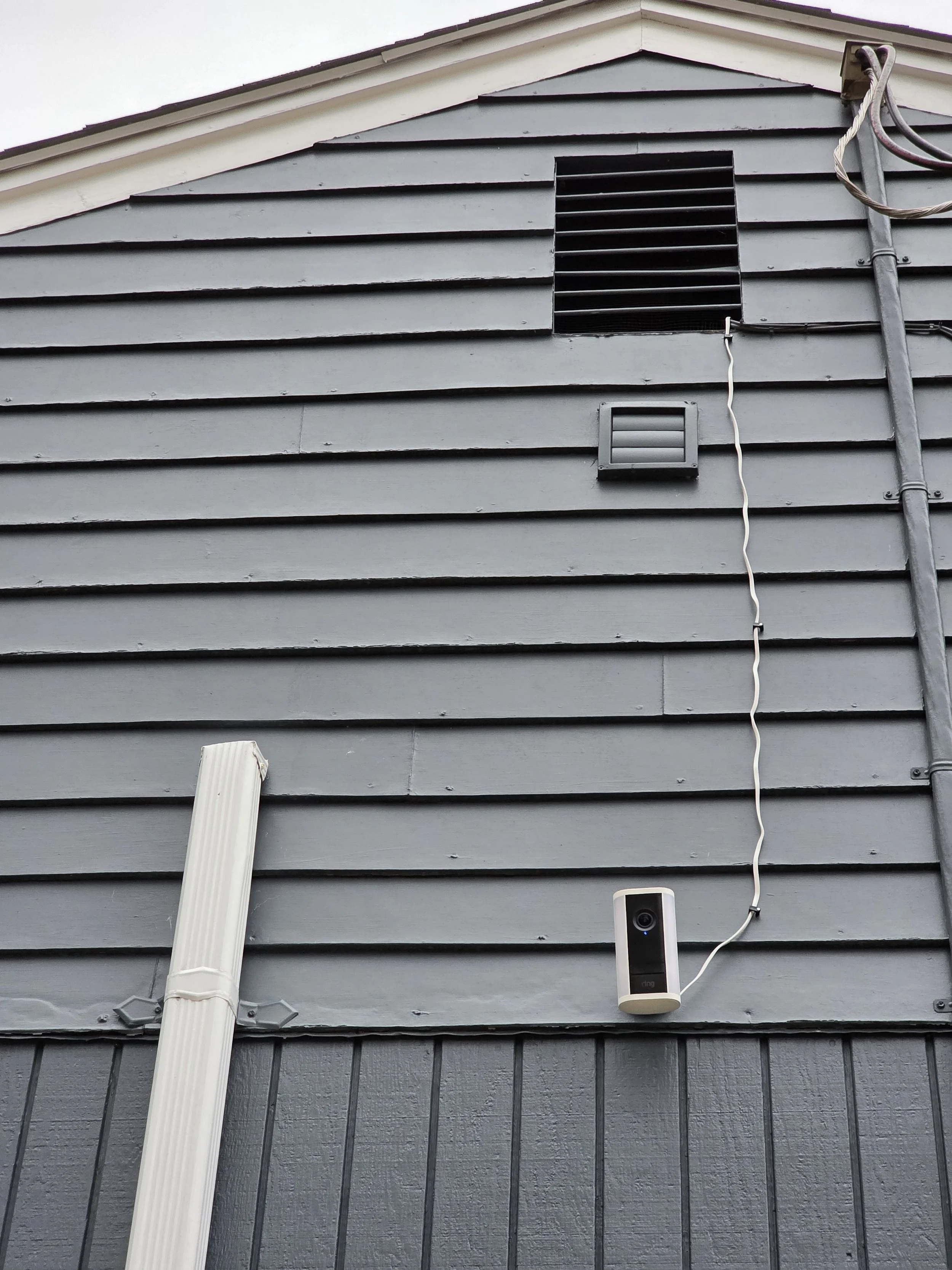 The side of a house with gray horizontal siding, a small ventilation vent, a black louvered vent, a white security camera, and electrical cables.