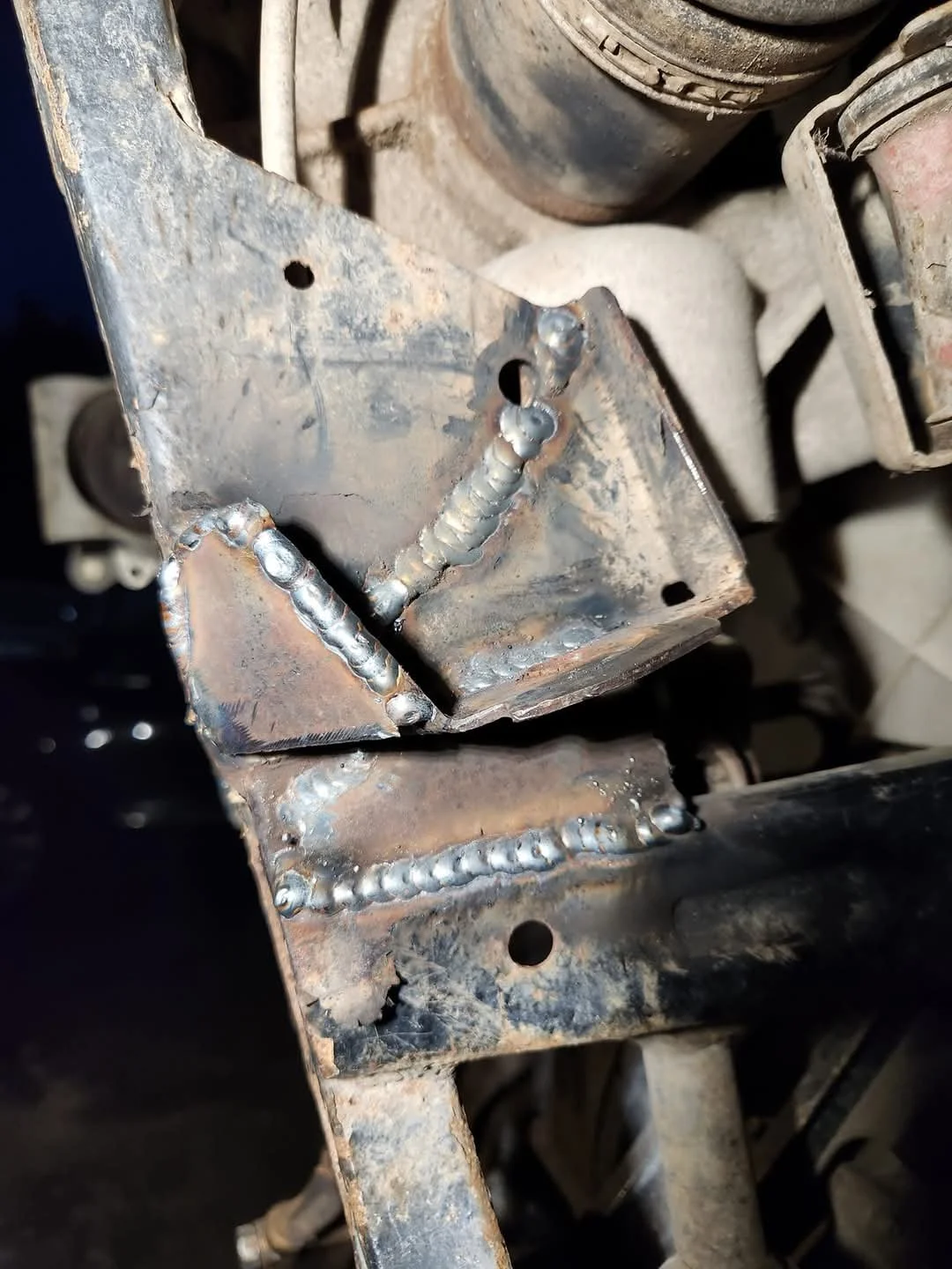 Close-up of a metal bracket with visible welds, attached to a larger metal structure and surrounded by dusty mechanical parts.