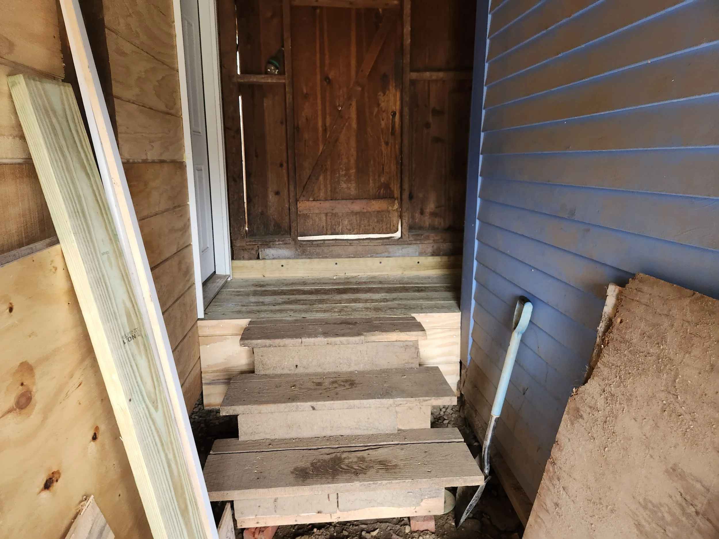 Wooden stairs leading to a door inside a partially constructed or renovated building with exposed wood and siding.